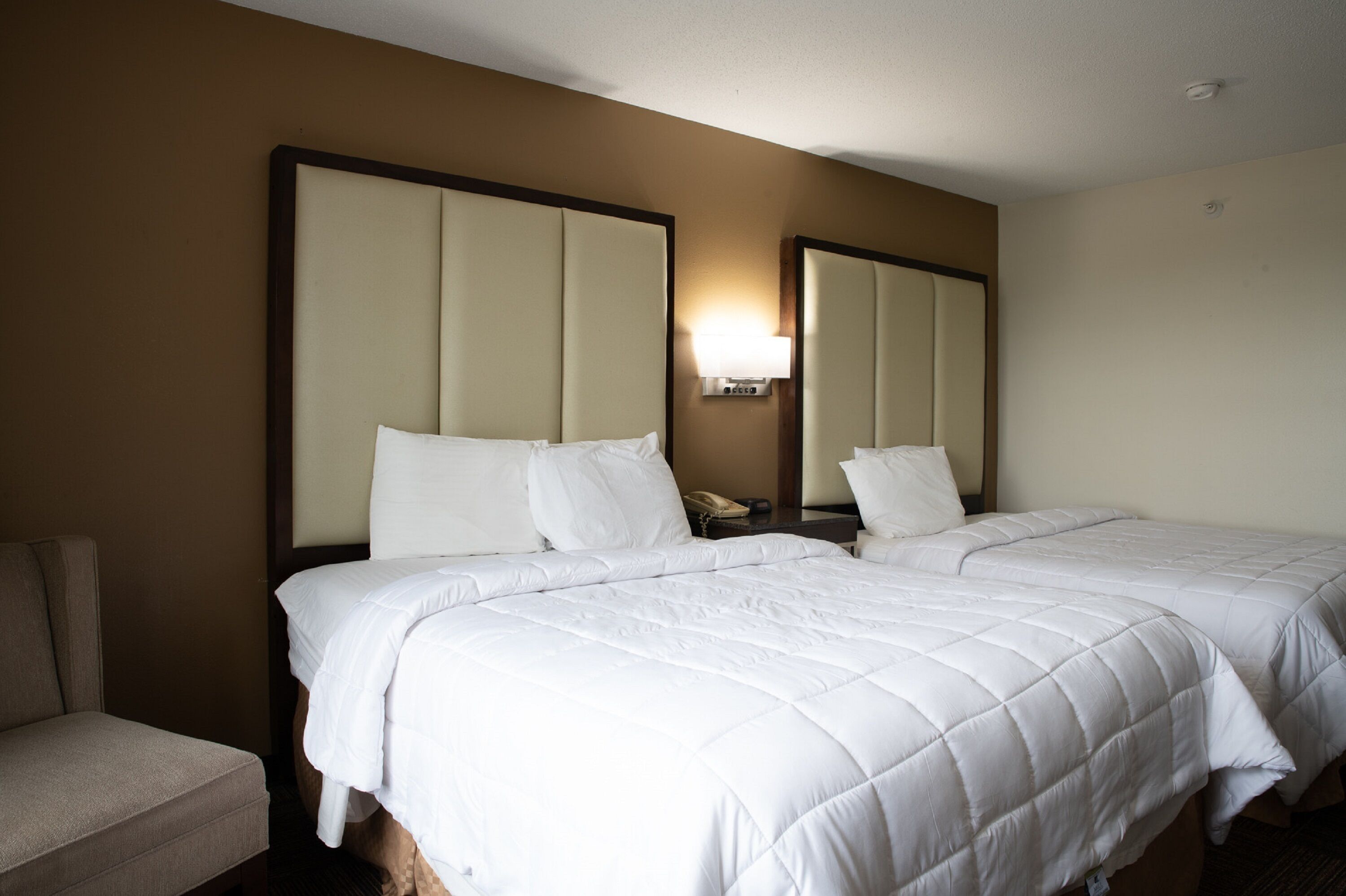 A hotel room with two beds, a nightstand, a lamp, and a chair.