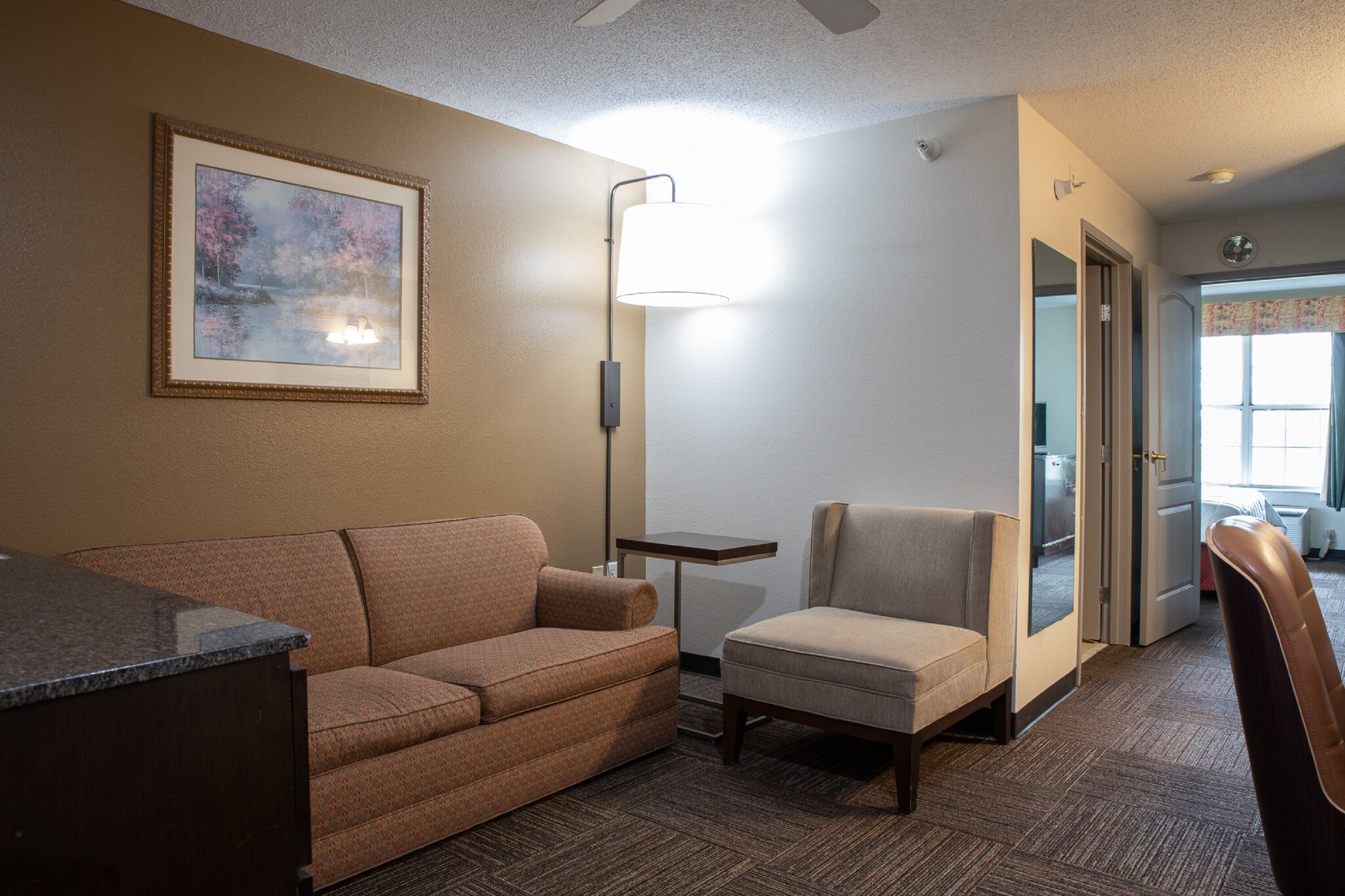 A hotel room with a sofa, armchair, and a desk. There is a framed picture on the wall and a lamp on a side table.