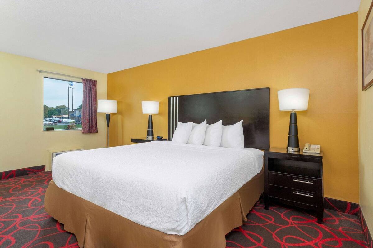Une chambre d’hôtel avec un grand lit, deux tables de chevet avec des lampes, une fenêtre donnant sur un parking et une porte.