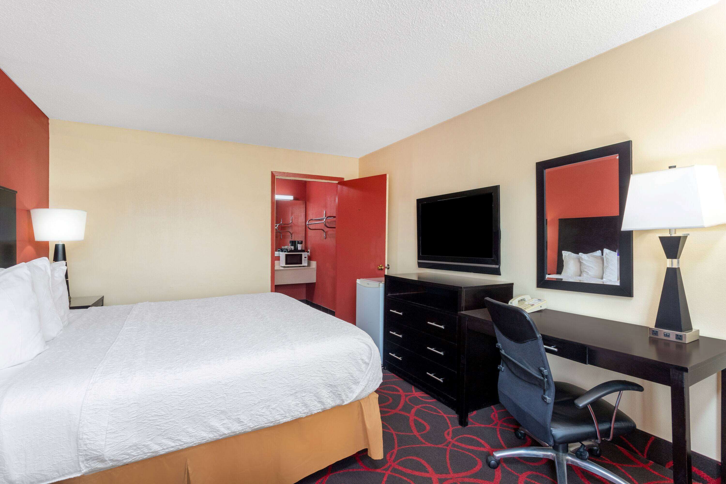A hotel room with a large bed, a desk with a chair, a flat-screen TV, a mirror, and a lamp.