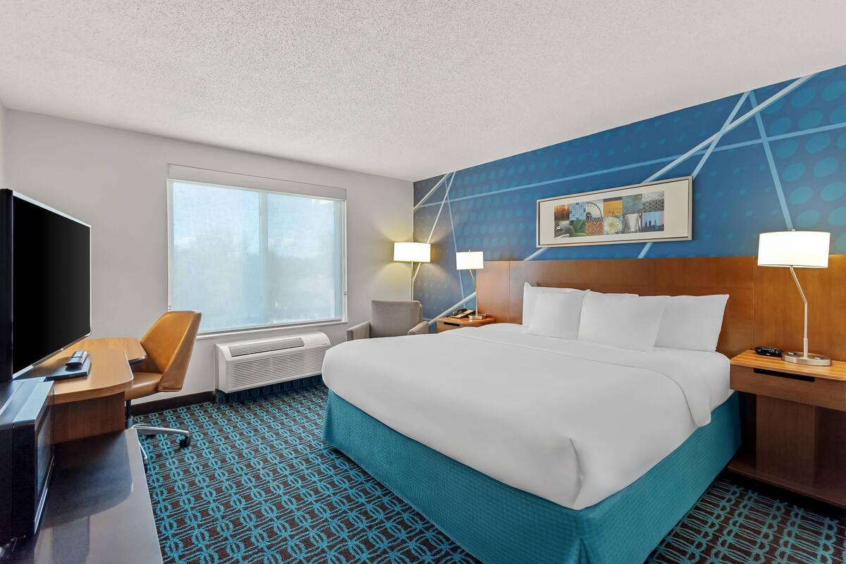 Habitación de hotel con una cama grande, un escritorio con televisor, una silla, un ventanal y una pared con un diseño geométrico.