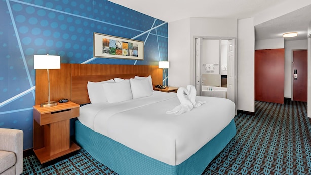 Quarto de hotel com uma cama grande, duas mesas de cabeceira com abajures, cabeceira de madeira, parede com padrão azul, um quadro emoldurado e uma porta que dá para um banheiro.