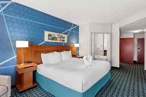 A hotel room with a large bed, two bedside tables with lamps, a wooden headboard, a blue patterned wall, a framed picture, and a door leading to a bathroom.