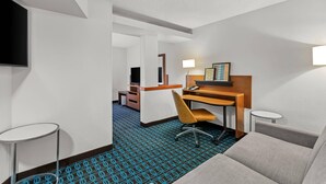 Quarto de hotel com sofá, escrivaninha com cadeira, televisão na parede, luminária e mesinha.