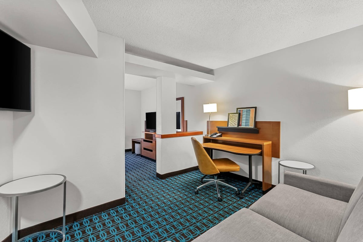 Habitación de hotel con sofá, escritorio con silla, televisor montado en la pared, lámpara y una mesita.