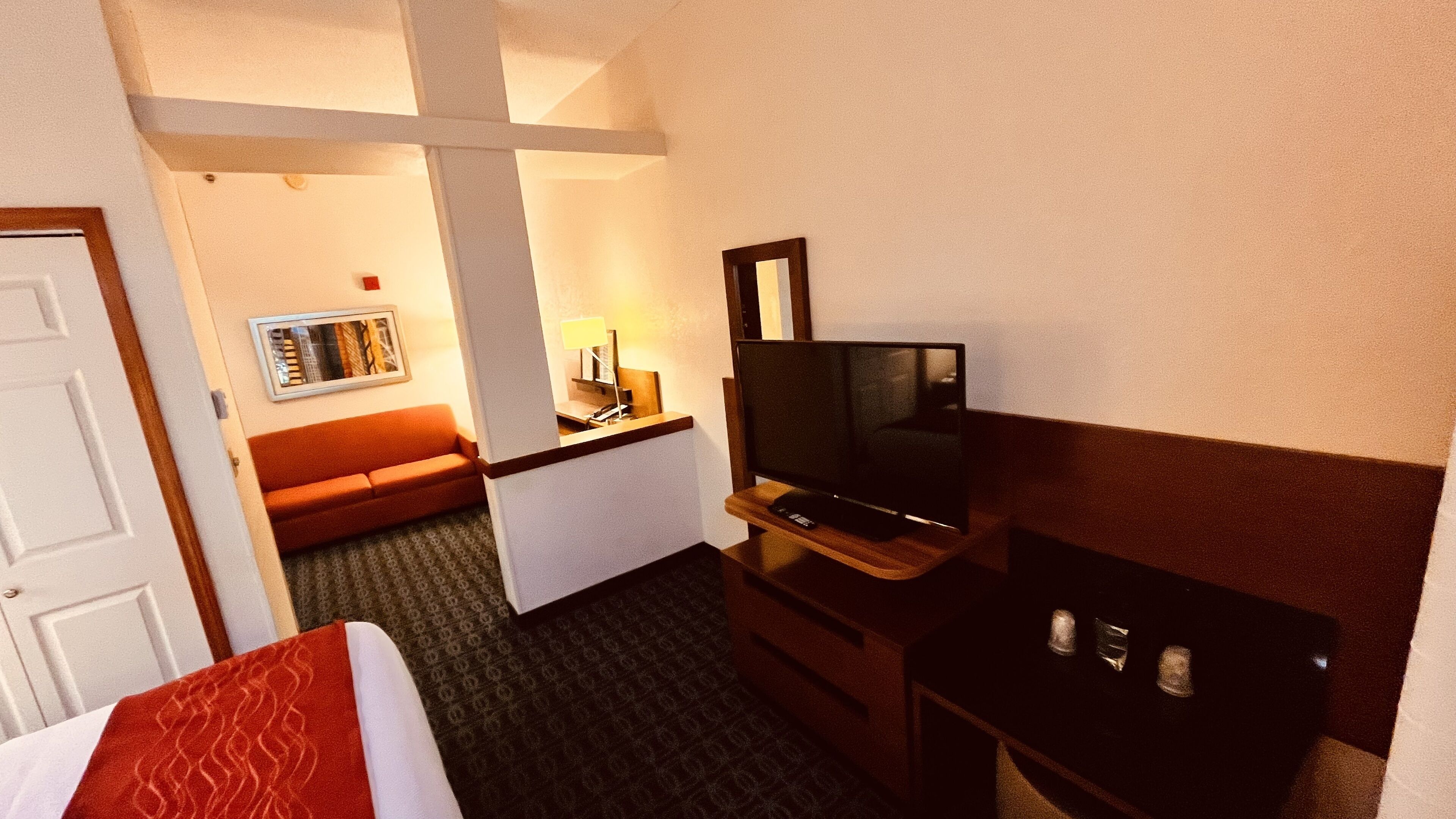 A hotel room with a bed, a TV, a desk, a chair, a sofa, and a small table with a lamp.
