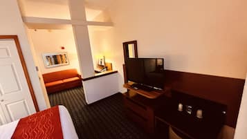 A hotel room with a bed, a TV, a desk, a chair, a sofa, and a small table with a lamp.
