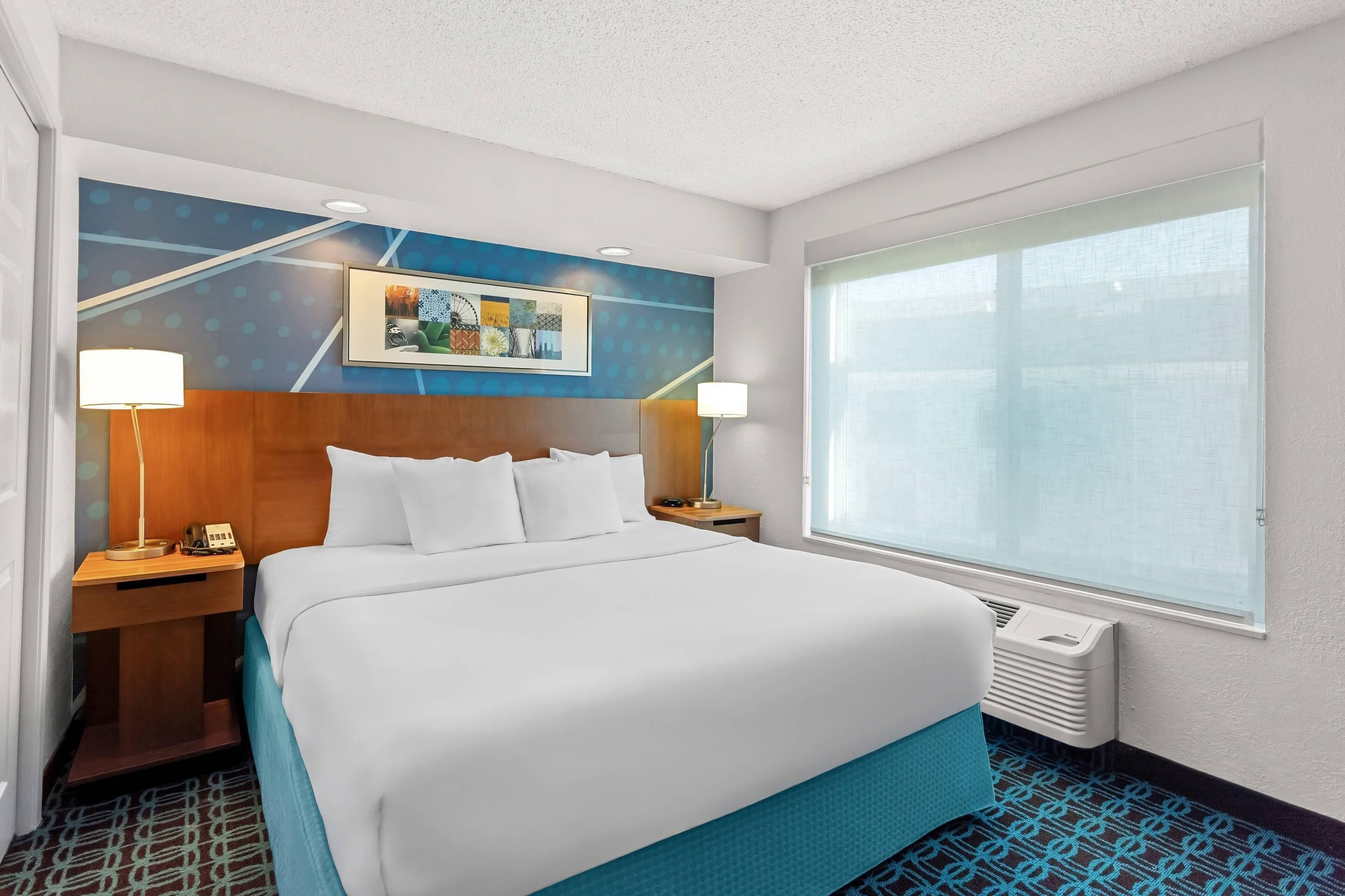 A hotel room with a large bed, two bedside tables, a window with blinds, and a wall with a geometric pattern.