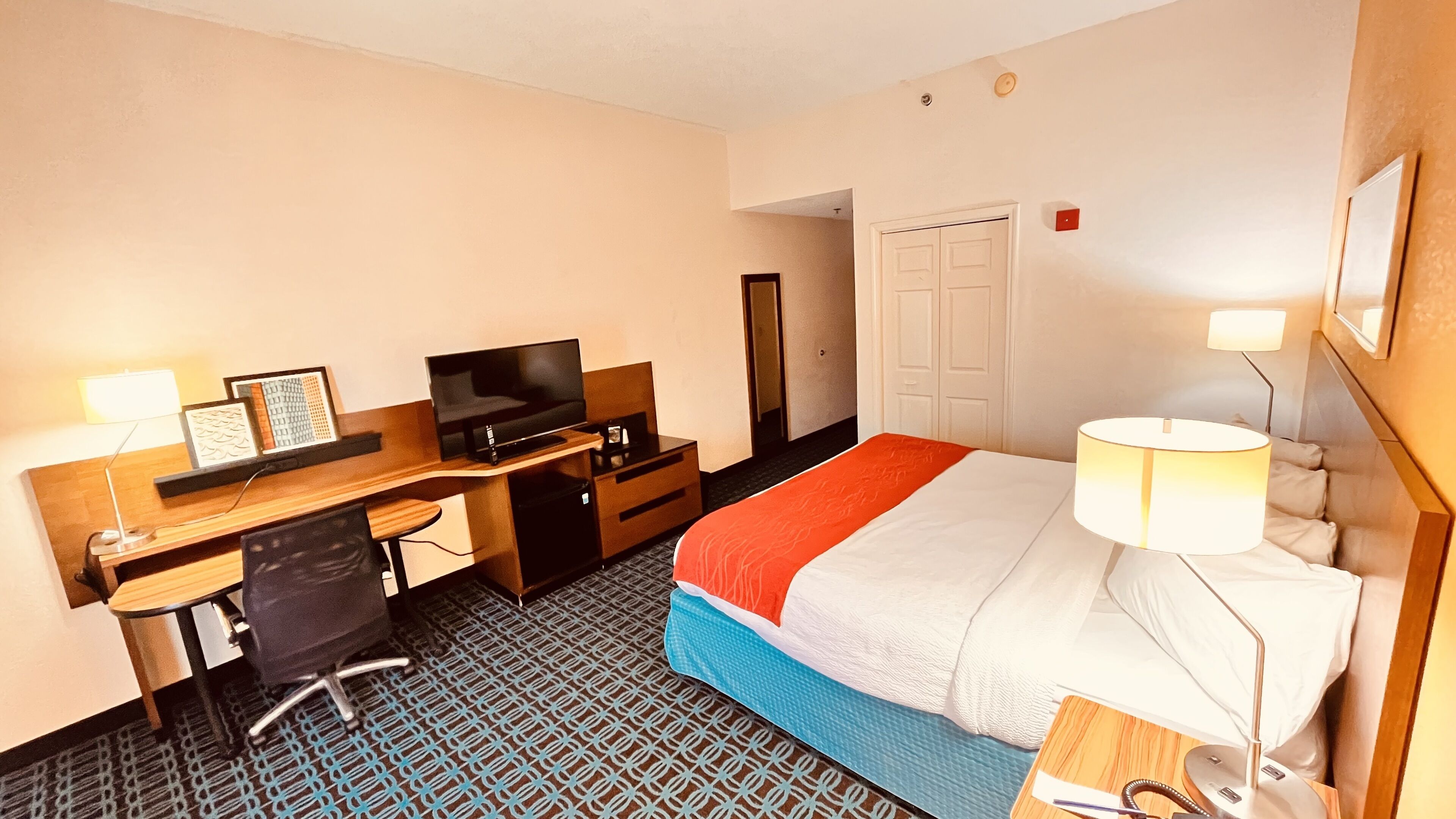 A hotel room with a bed, desk, TV, chair, and a lamp.