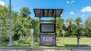 Uma estação de carregamento para veículos elétricos alimentada por energia solar, com uma placa indicando "EOS CHARGE" e um endereço de site.