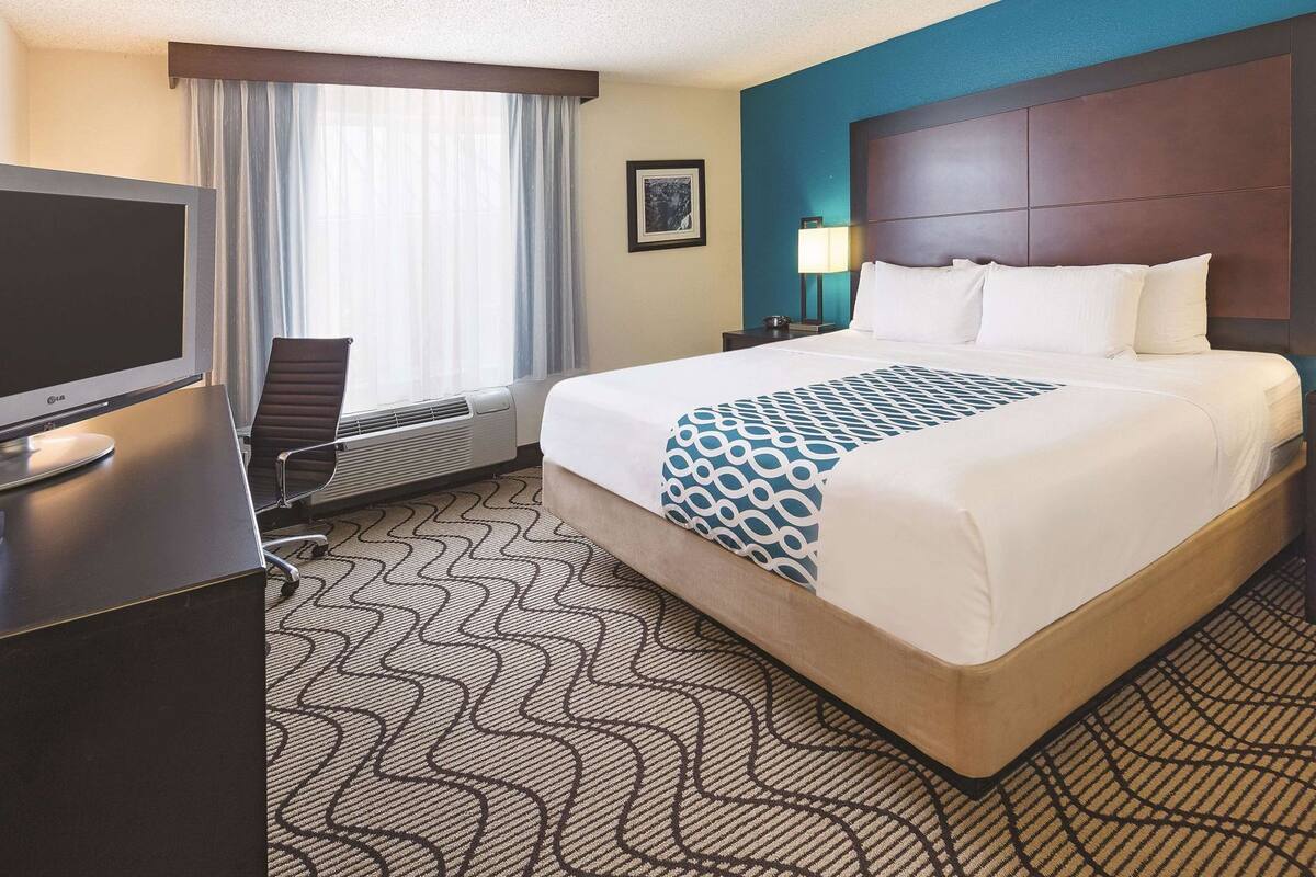 A hotel room with a bed, a desk with a TV, a chair, a window with curtains, and a nightstand with a lamp.