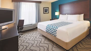 A hotel room with a bed, a desk with a computer, a chair, a window with curtains, and a nightstand with a lamp.