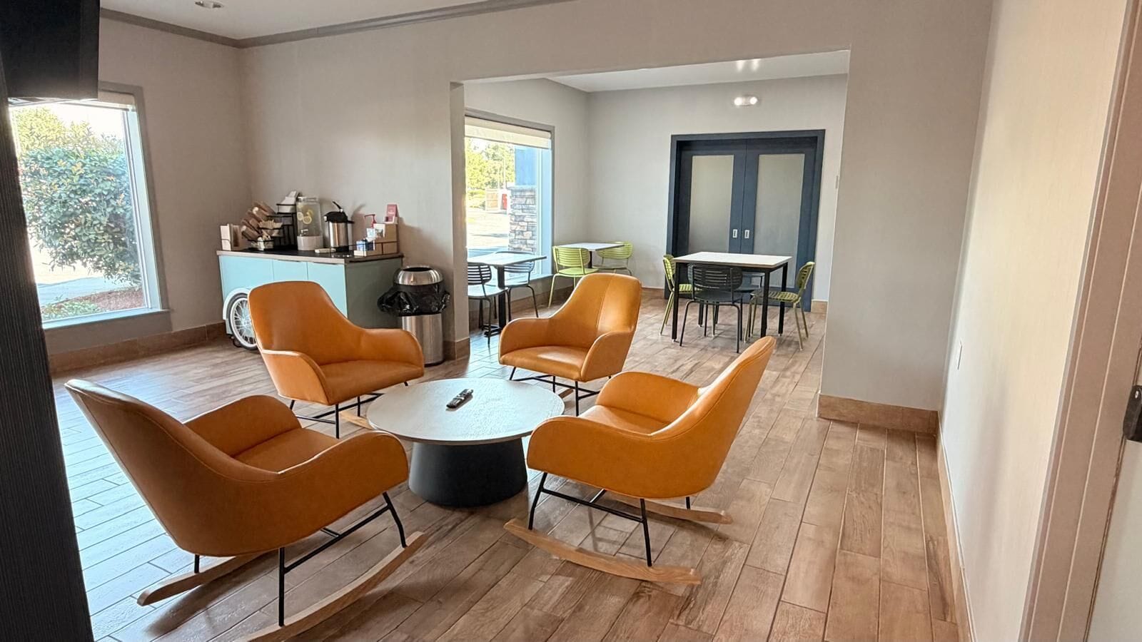 A modern interior with a round table surrounded by orange chairs, a kitchenette area, and a view through a glass door.