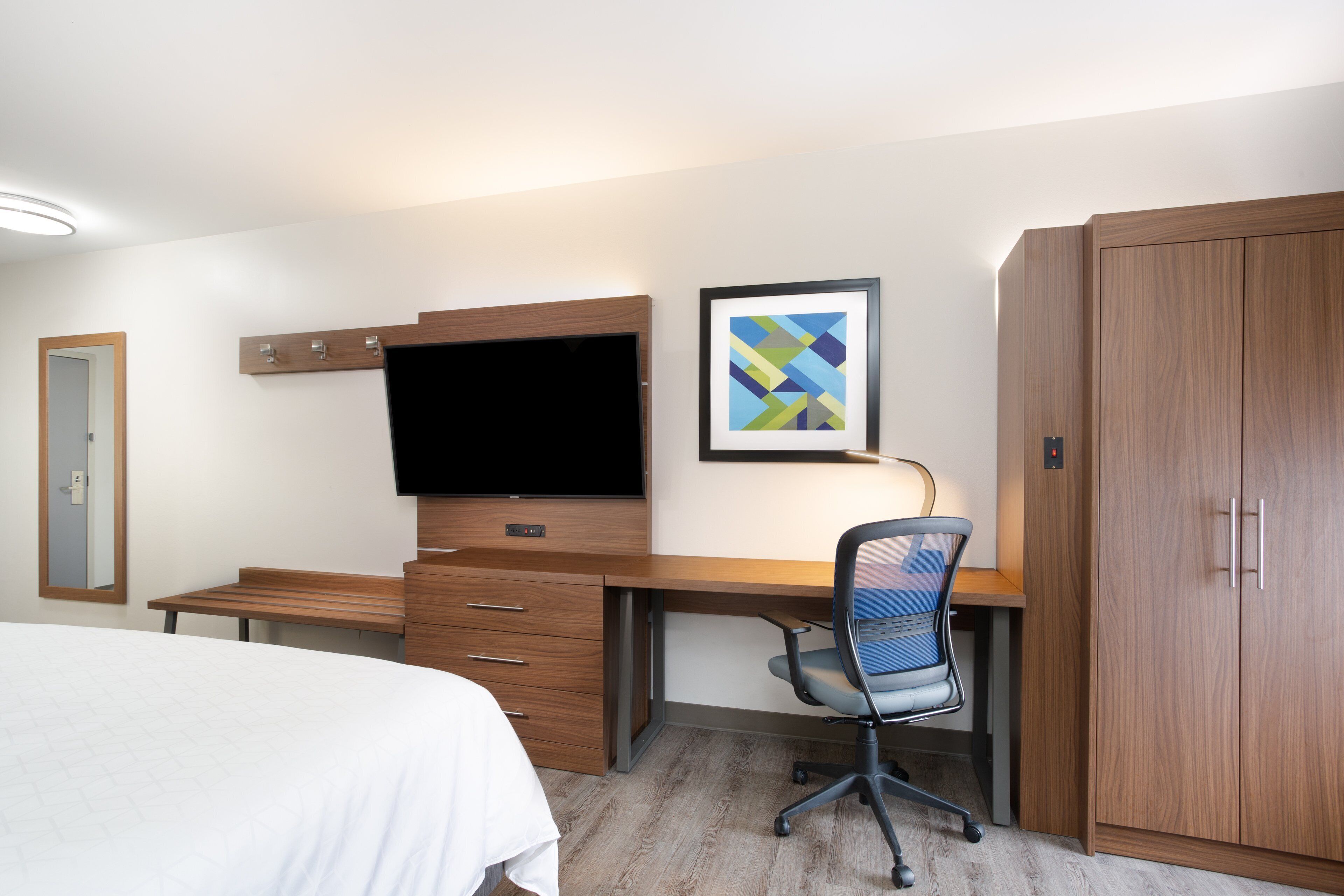 Ein Hotelzimmer mit einem Bett, einem Schreibtisch mit Stuhl, einem Fernseher, einem Kleiderschrank und einem gerahmten Bild an der Wand.