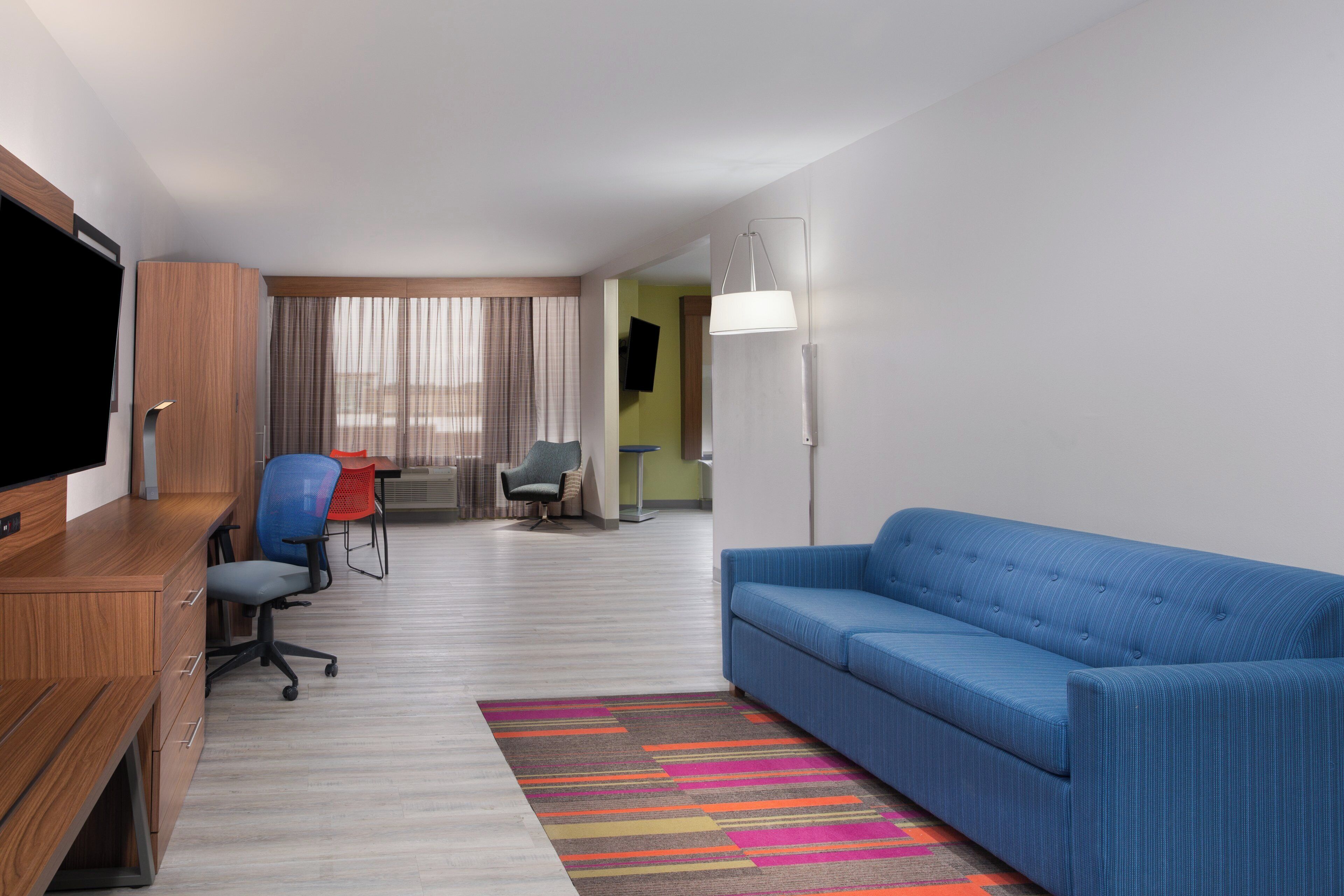 A hotel room with a television, a blue sofa, a desk with a chair, and a colorful rug.