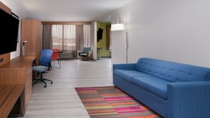 A hotel room with a wooden desk, a blue sofa, a colorful striped rug, a television, and a flat-screen TV mounted on the wall.