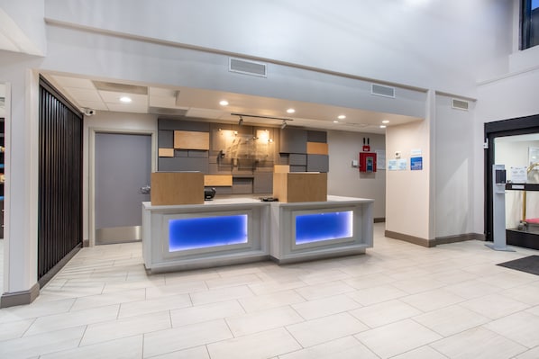 A reception area with a modern design, featuring a counter with illuminated blue panels, a wooden panel wall, and a ceiling with recessed lighting.
