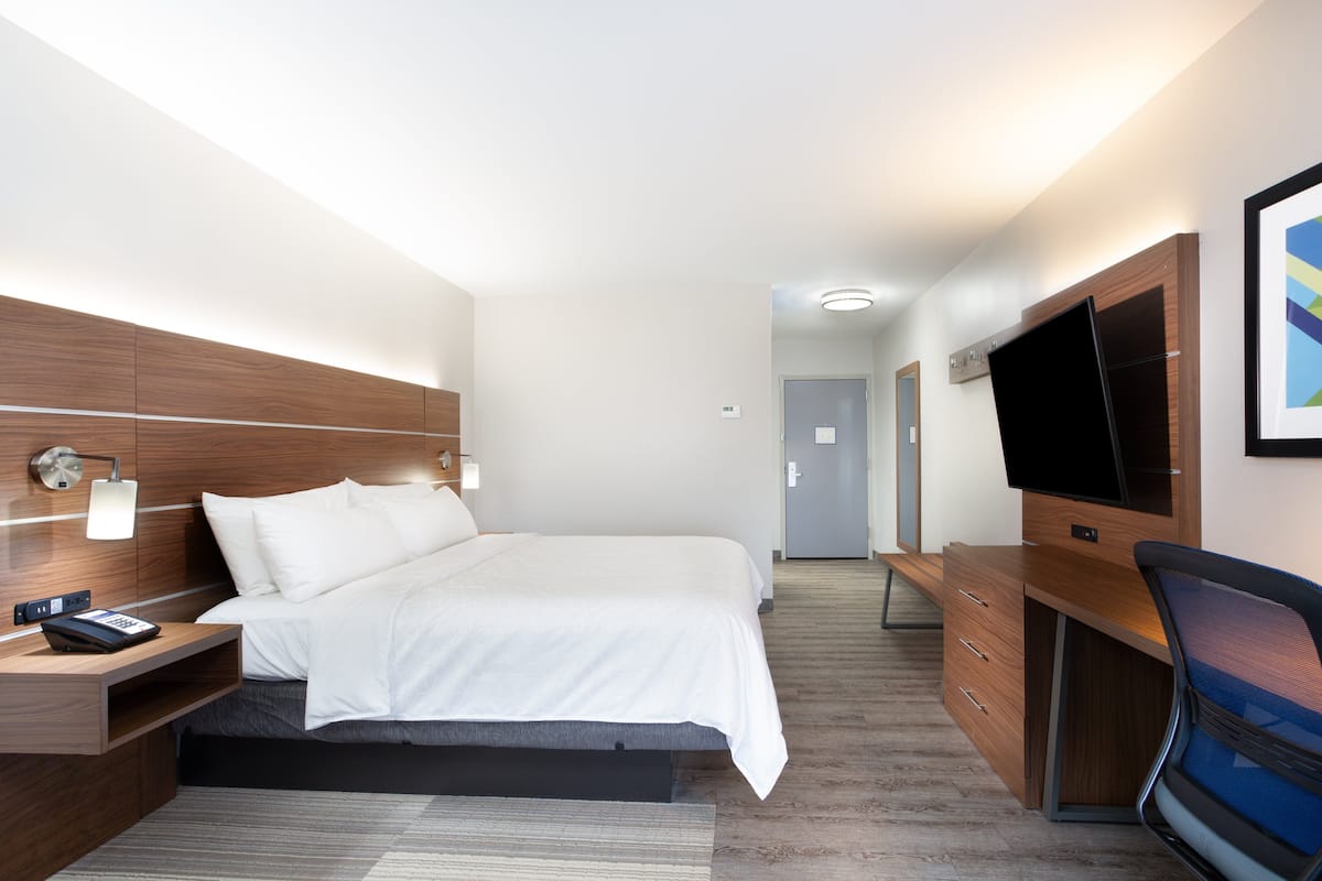 A modern hotel room with a large bed, bedside table with a phone, a desk with a TV, and a chair.