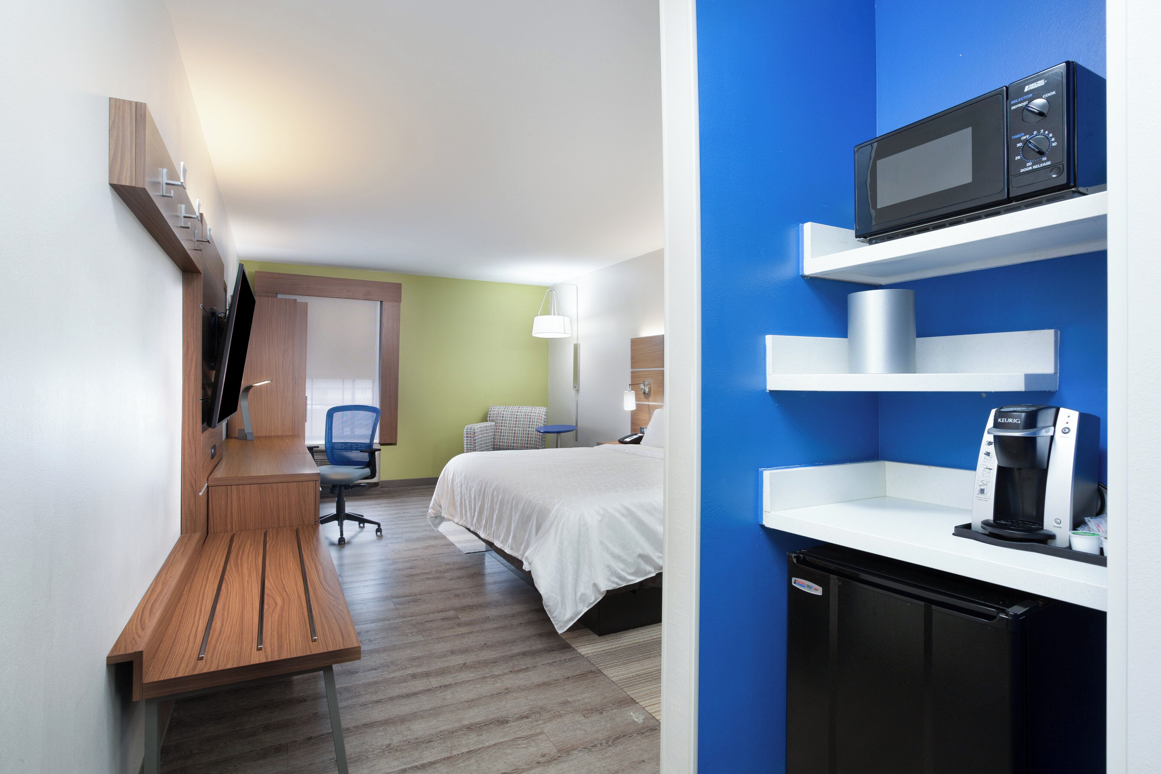 A hotel room with a desk, bed, microwave, coffee maker, and a small refrigerator.