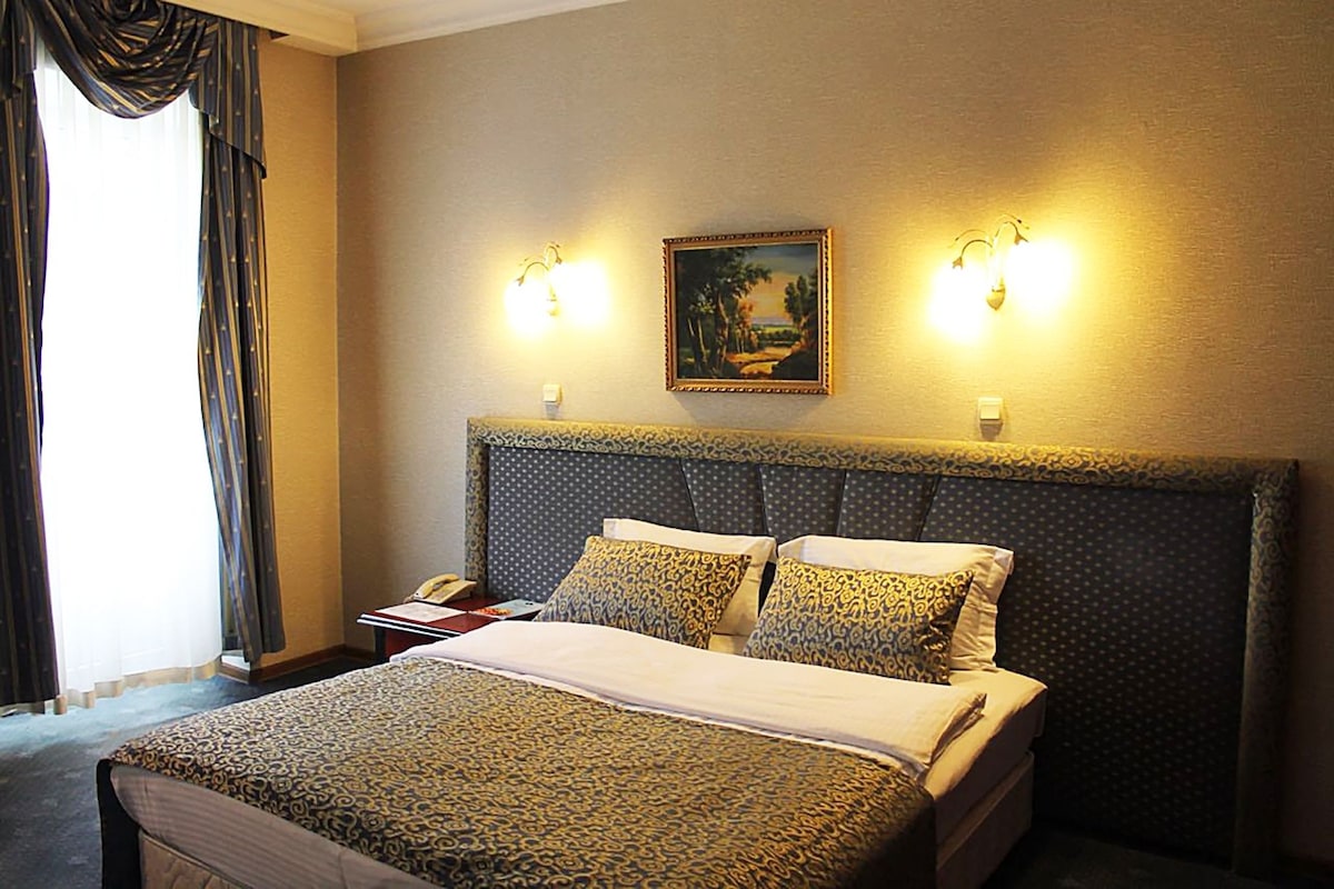 A hotel room with a large bed, two bedside lamps, a painting on the wall, and a window with curtains.