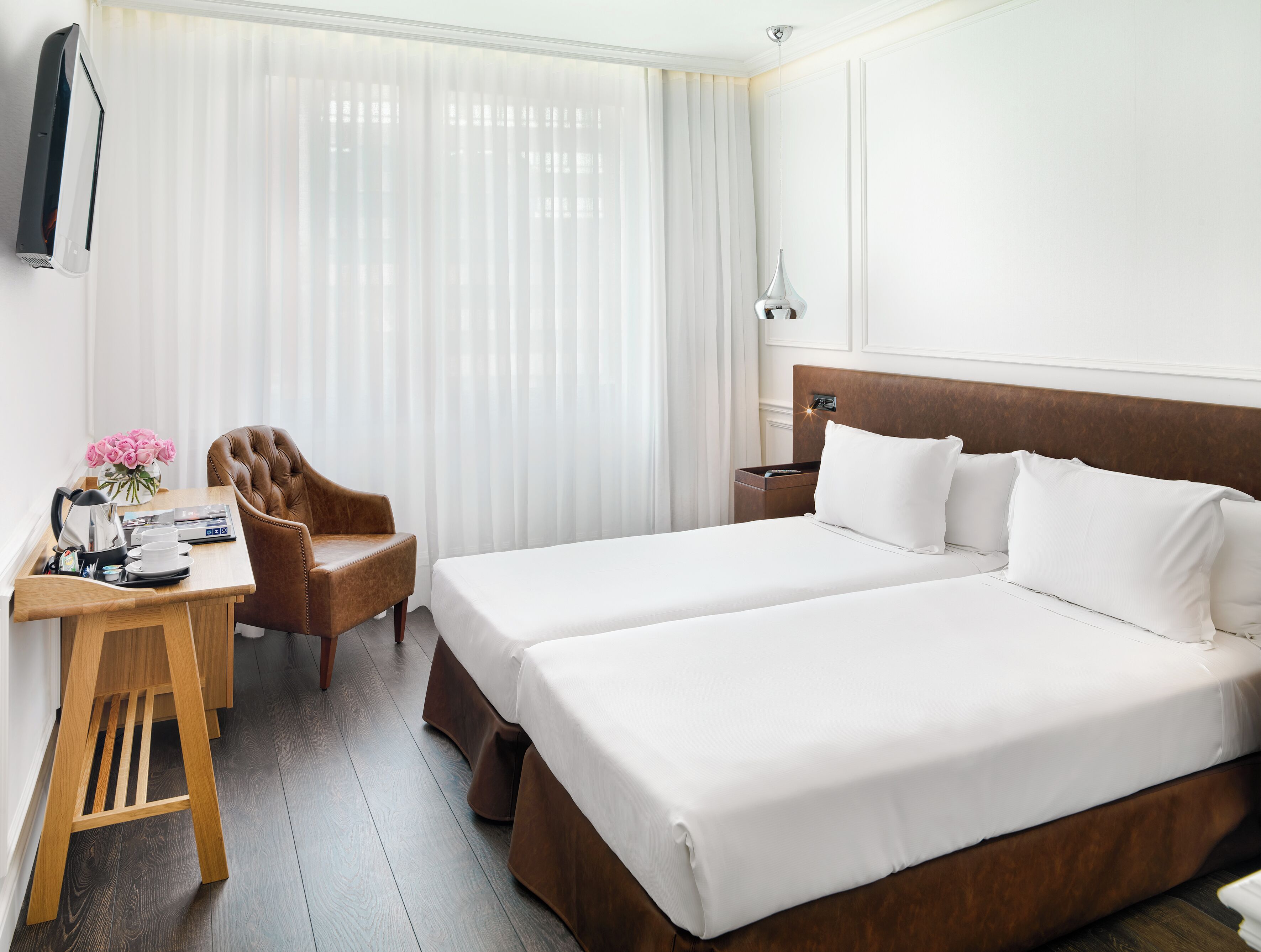 Une chambre d’hôtel avec un lit double, un bureau avec une chaise, une table de chevet et une grande fenêtre avec des rideaux légers.