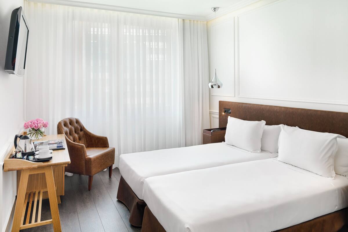 Une chambre d’hôtel avec un lit double, un bureau avec une chaise, une table de chevet et une grande fenêtre avec des rideaux légers.