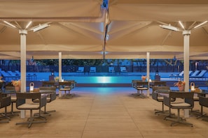 Ein Außenpoolbereich mit Liegestühlen, Tischen, einem gefliesten Boden und einem Pool im Hintergrund.