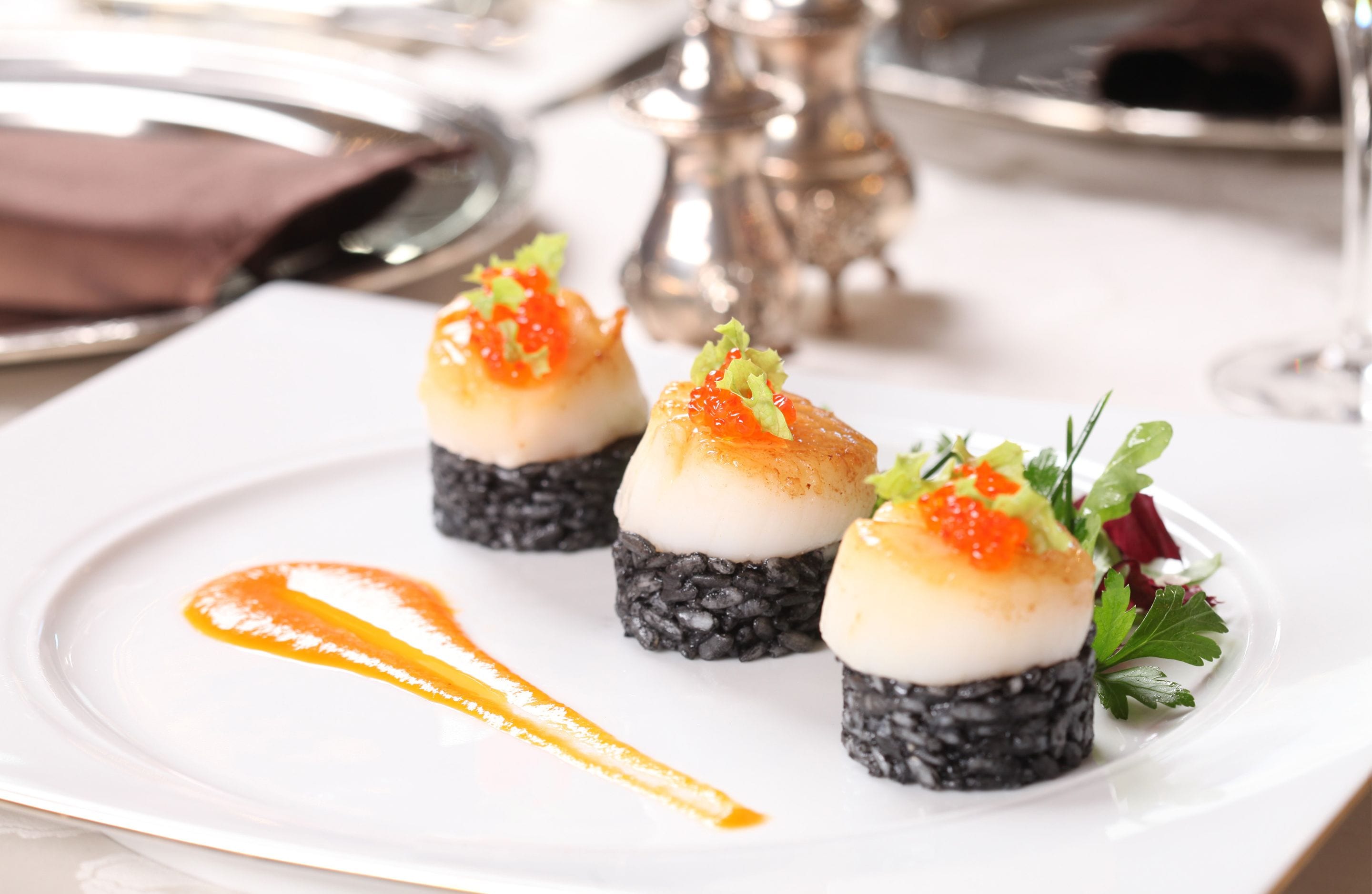Valkoinen lautanen, jossa on kolme sushikappaletta, päälle on laitettu kaviaaria ja koristeltu vihreillä kasviksilla, ja annoksen päällä on kastetta.