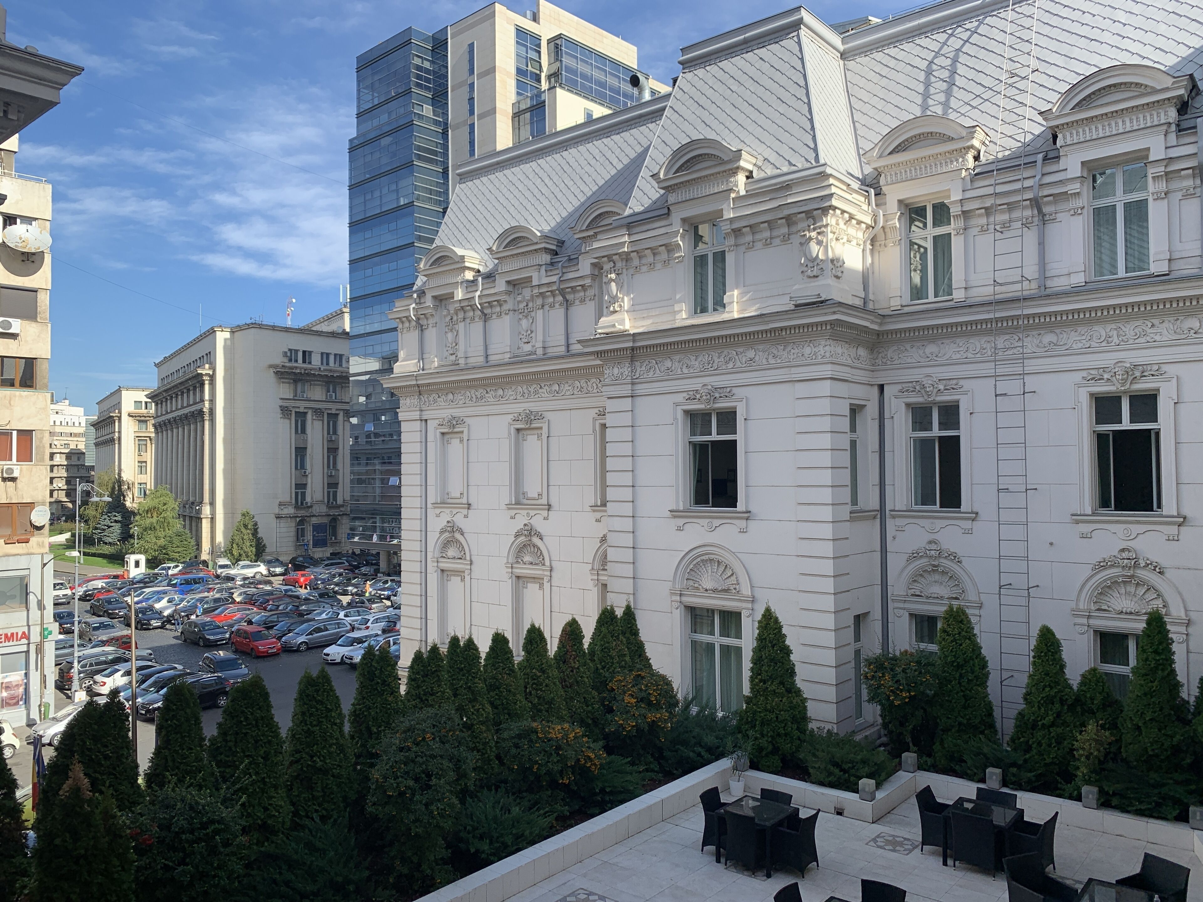 A historic building with classical architecture, a courtyard with trees, and a parking lot in the background.