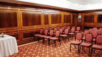 Un salón de conferencias con filas de sillas, una mesa blanca con una botella de agua y una alfombra estampada.