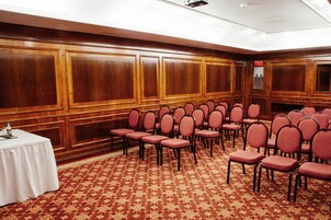 Un salón de conferencias con filas de sillas, una mesa blanca con una botella de agua y una alfombra estampada.