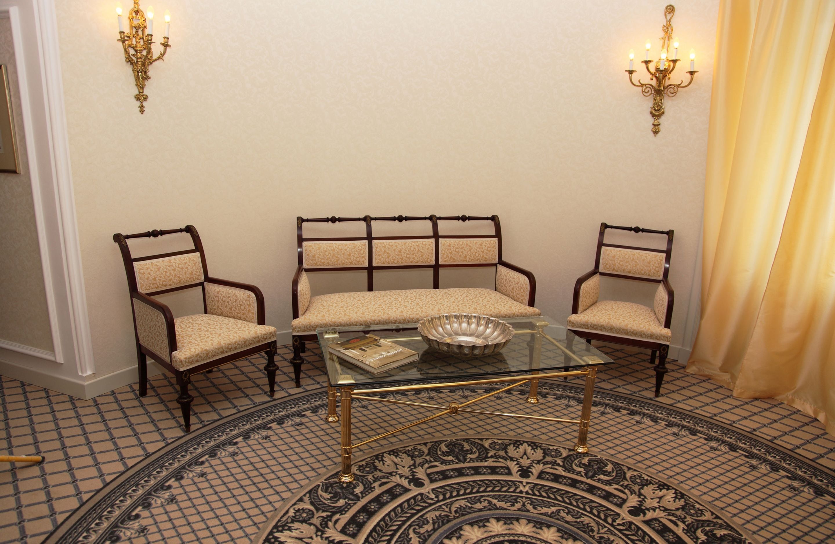 A room with a glass coffee table, three upholstered chairs, a patterned rug, and wall sconces.