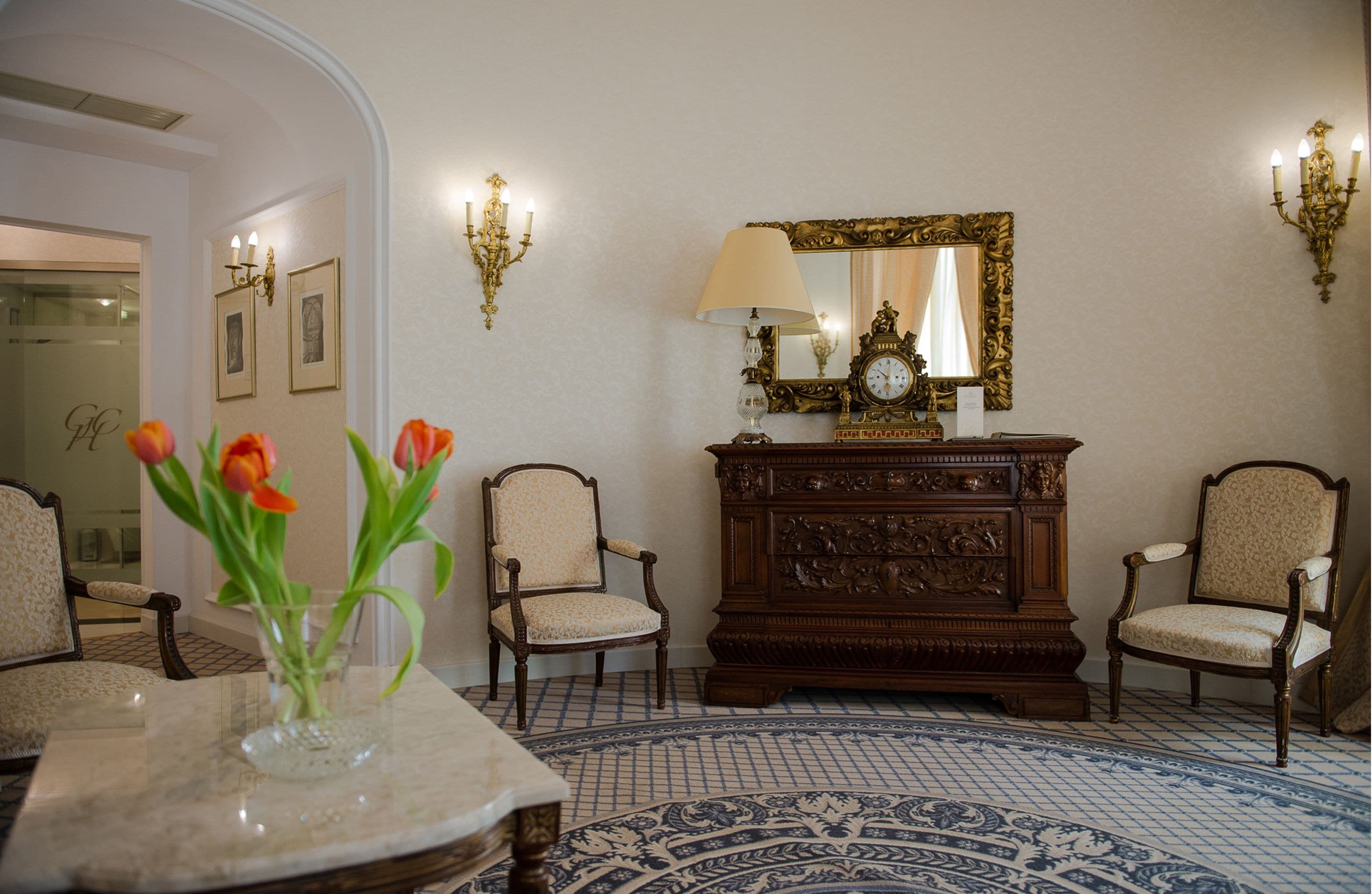 A room with two chairs, a table with a vase of flowers, a lamp, a mirror, and a patterned floor.