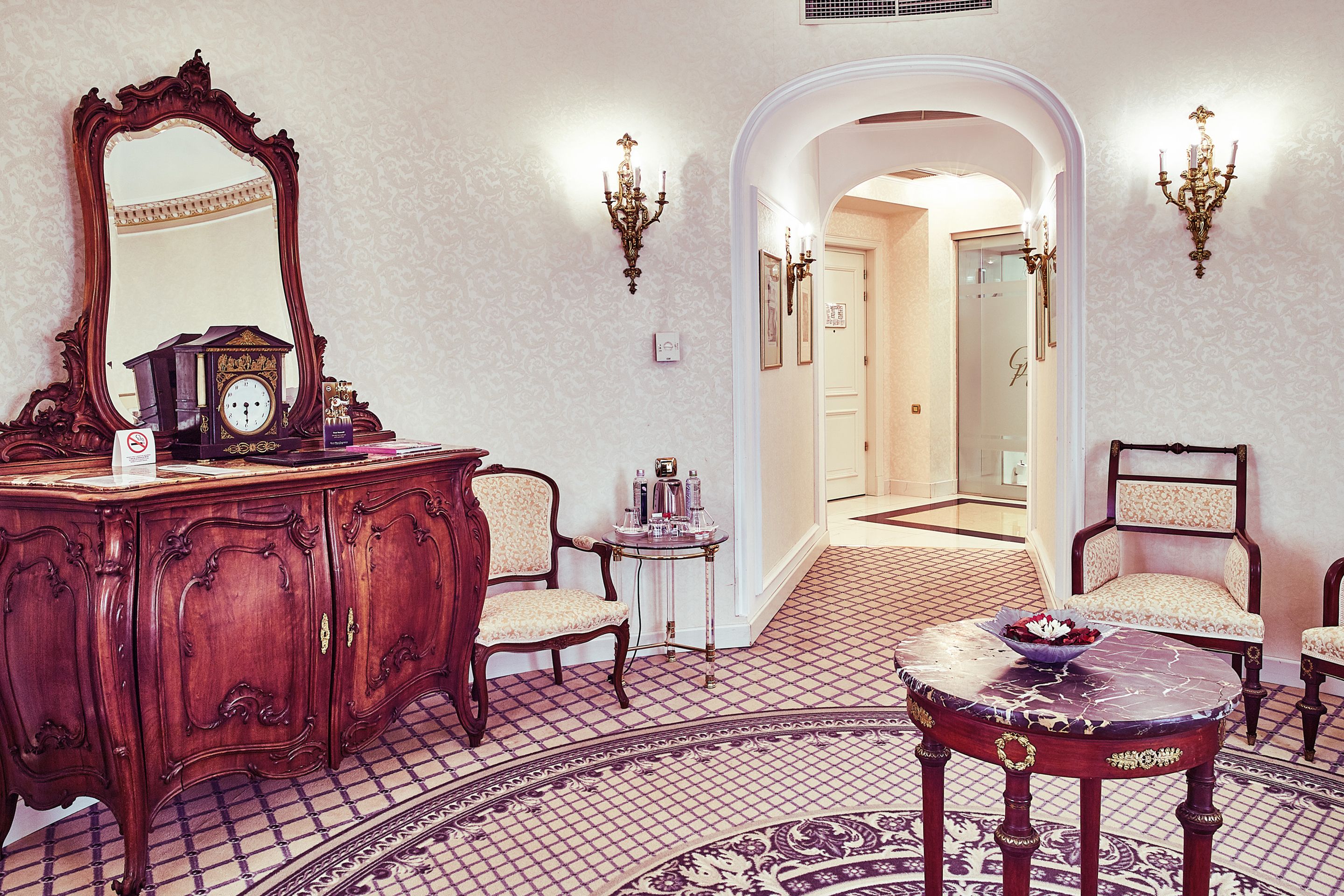 A room with a wooden dresser, a round table with a bowl, two chairs, a large mirror, and a patterned floor.