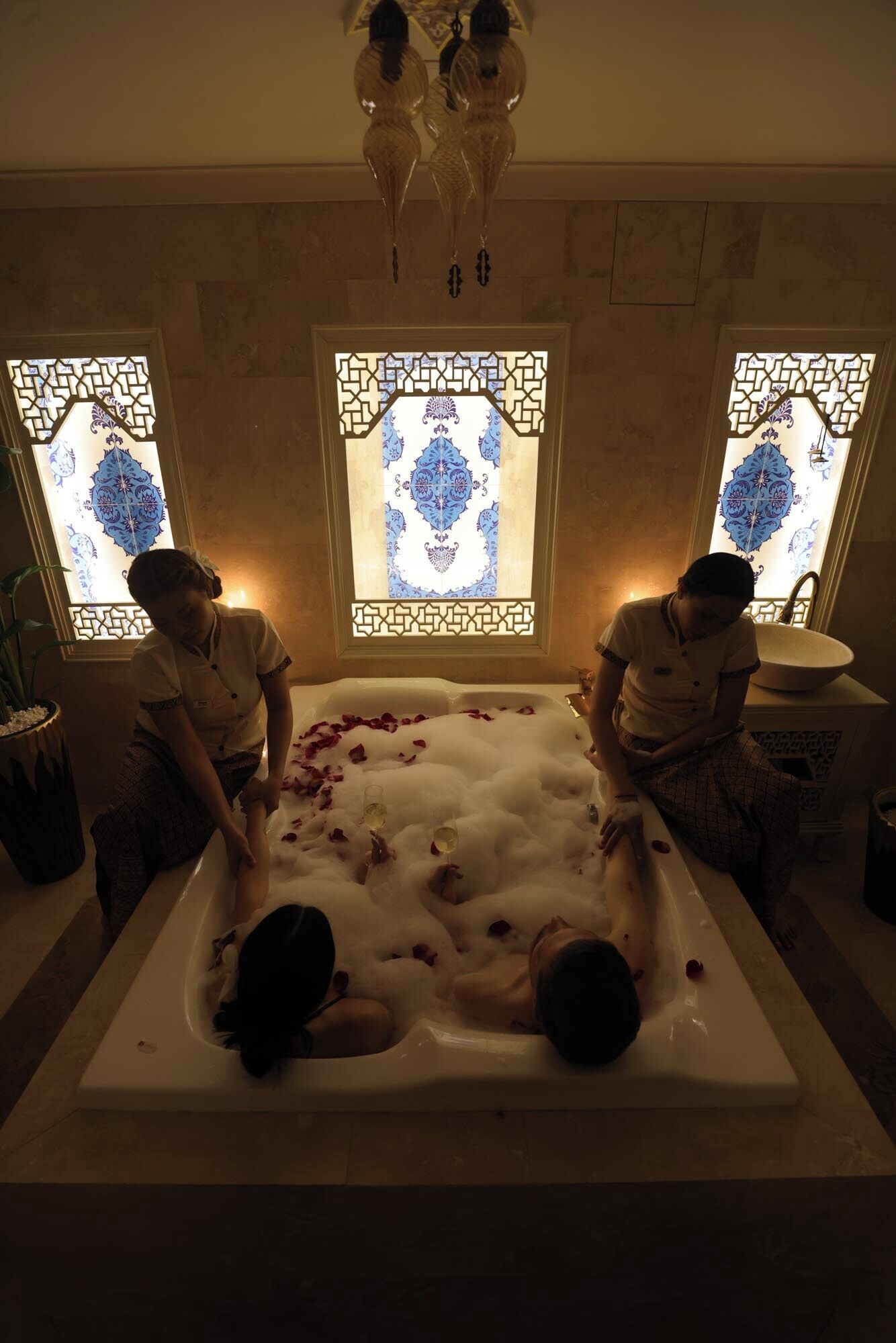 Una bañera llena de burbujas y pétalos de rosa, con dos asistentes.