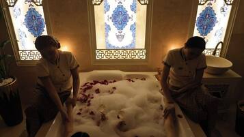 Una bañera llena de burbujas y pétalos de rosa, con dos asistentes.