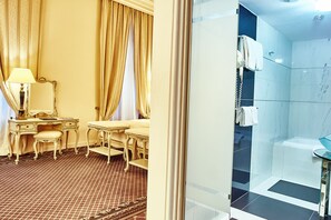 Una habitación con escritorio, silla y tocador con espejo. Incluye un baño con bañera, ducha con paredes de vidrio y retrete.