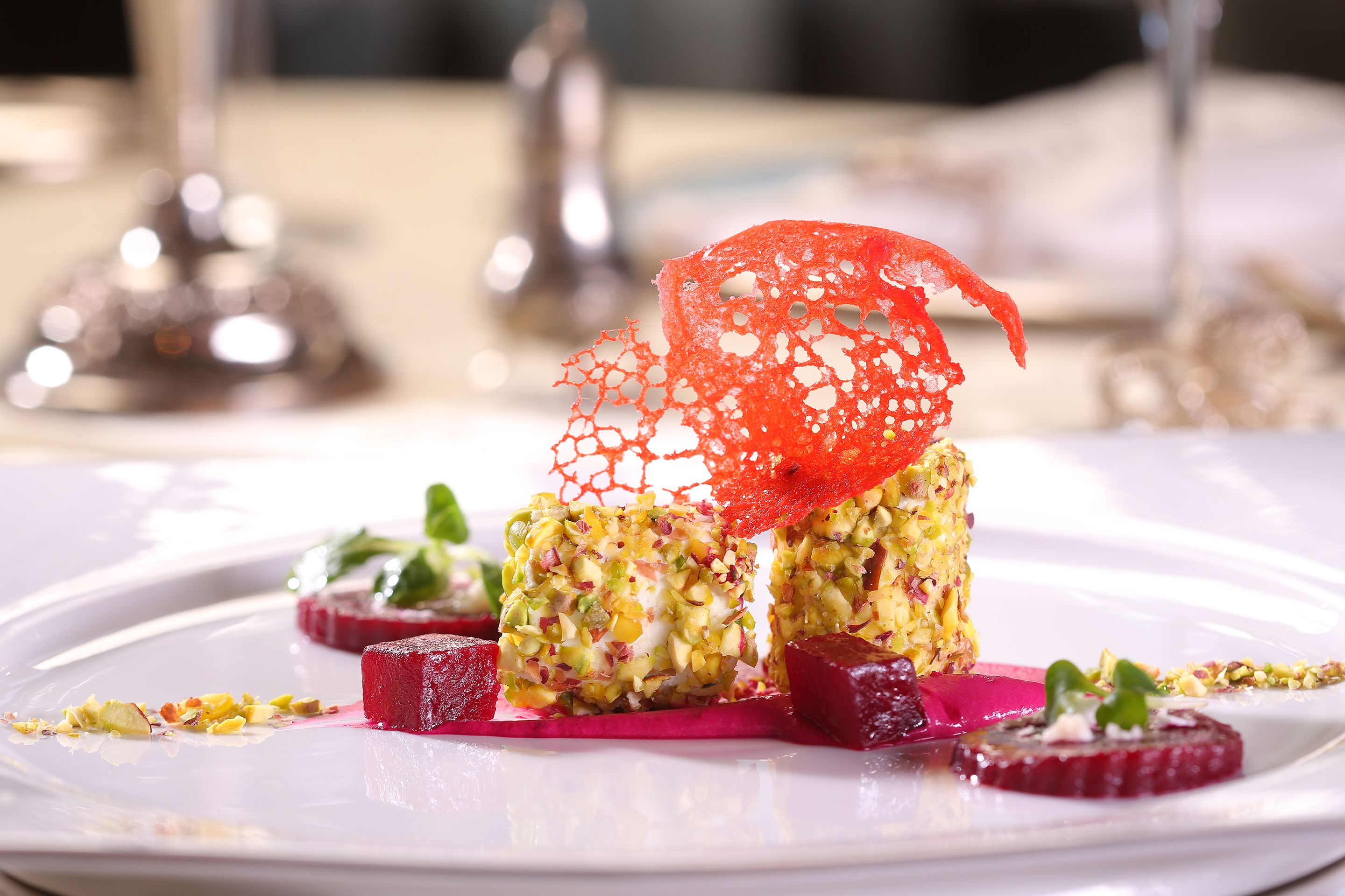 A dessert plate with a red net-like structure on top, surrounded by various garnishes and a white plate.