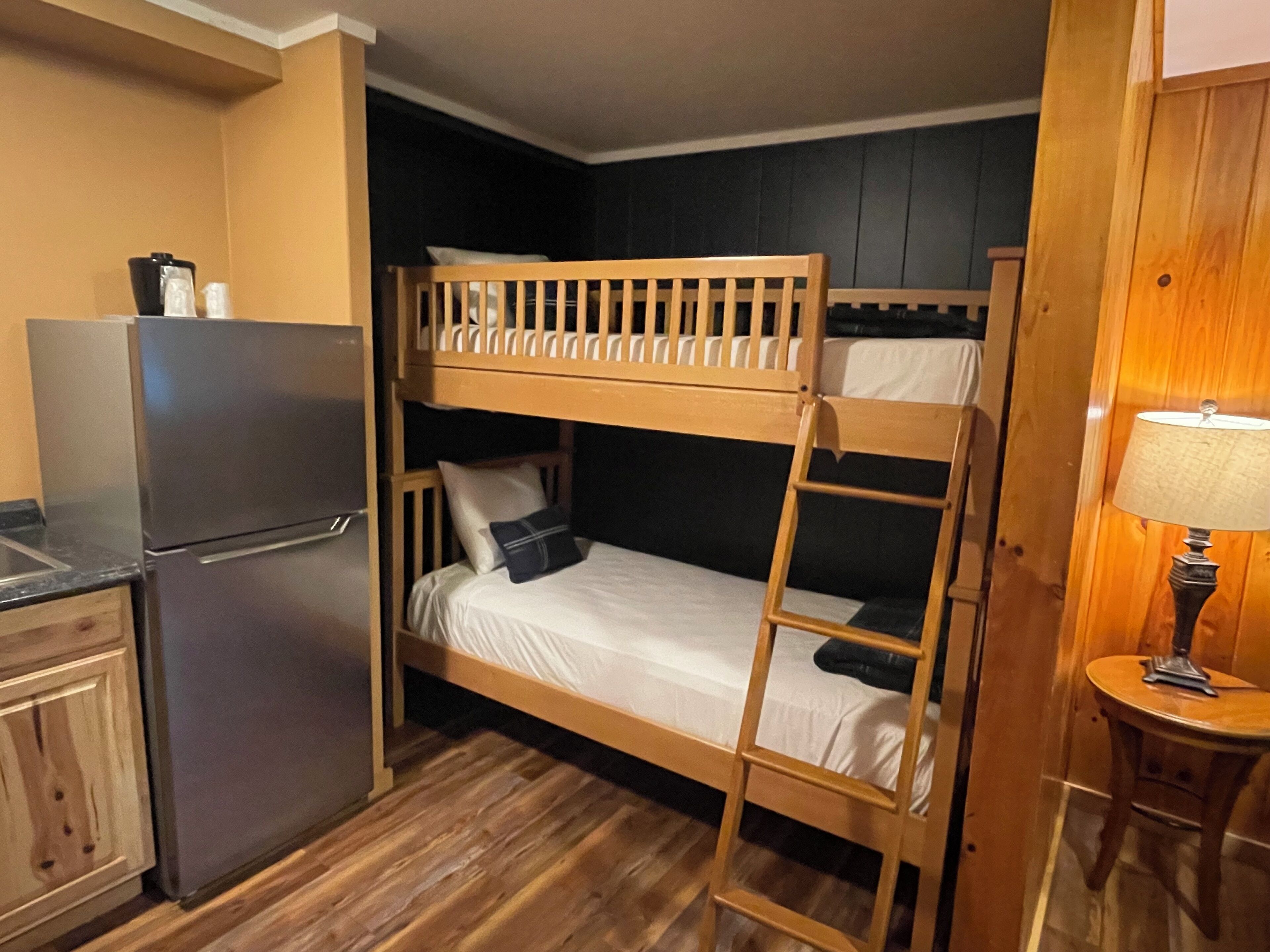 A bunk bed room with a refrigerator and a small table with a lamp.