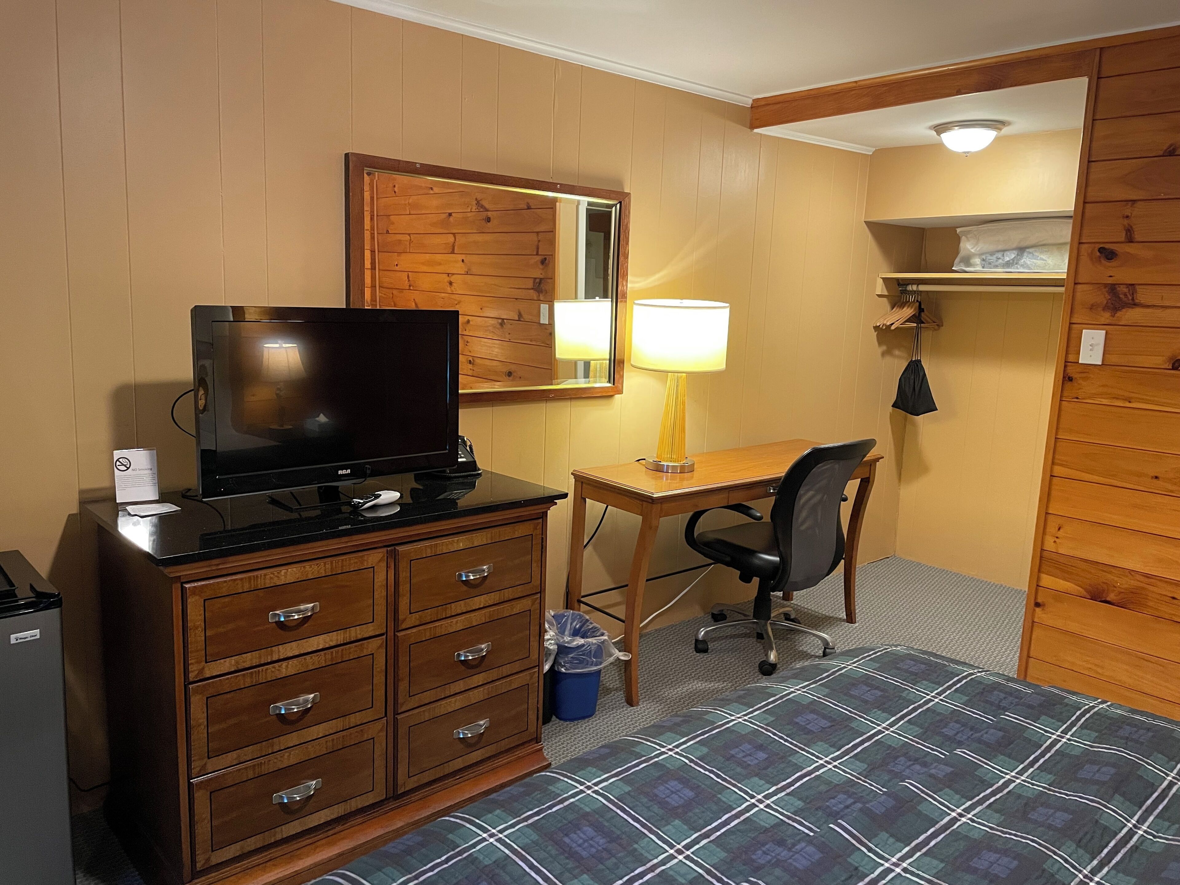 A compact hotel room with a wooden desk, a television, a lamp, a chair, a small trash bin, and a bed with a plaid bedspread.