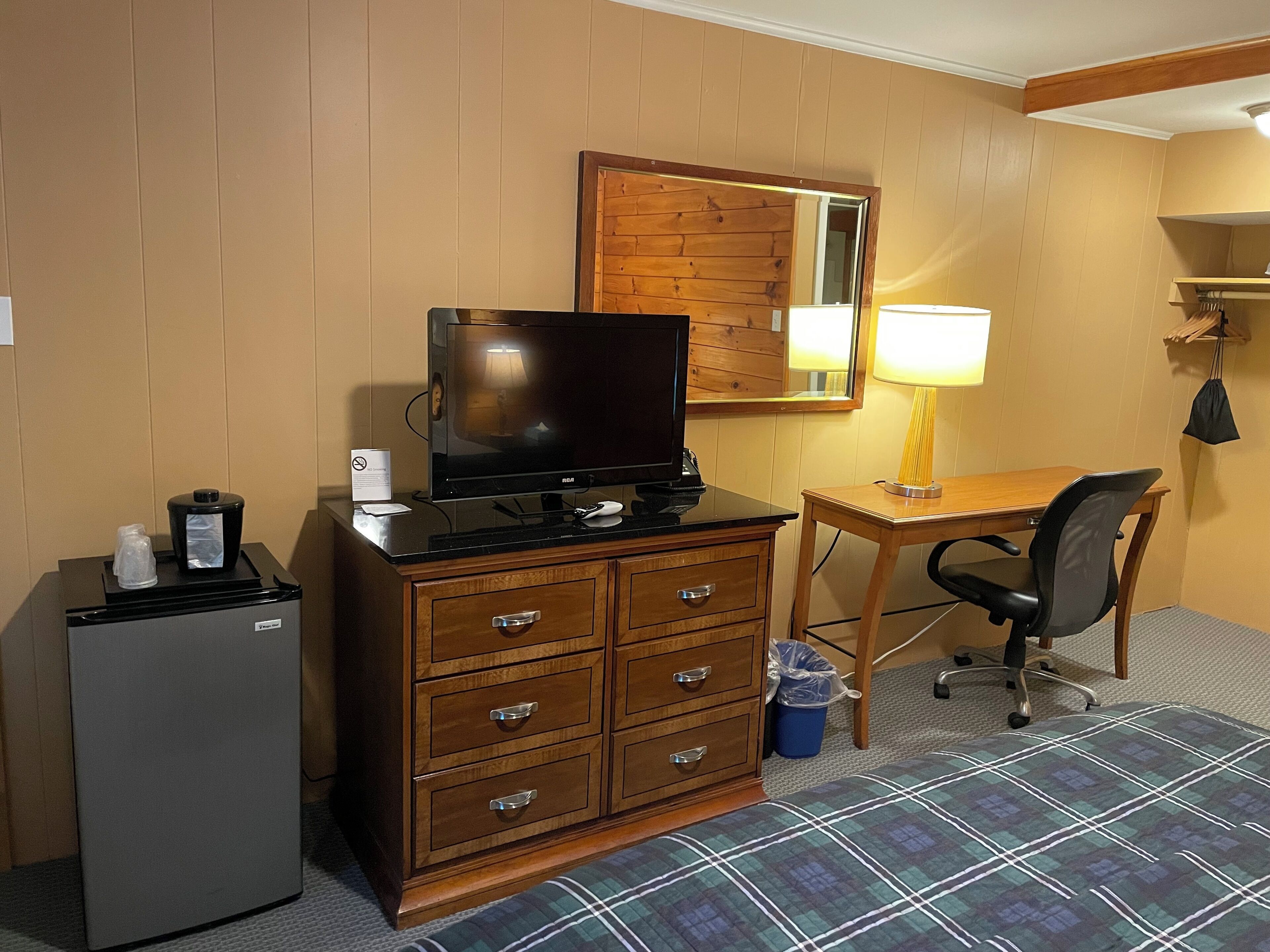 A hotel room with a wooden dresser, a television, a desk with a lamp, a chair, a small refrigerator, and a trash bin.