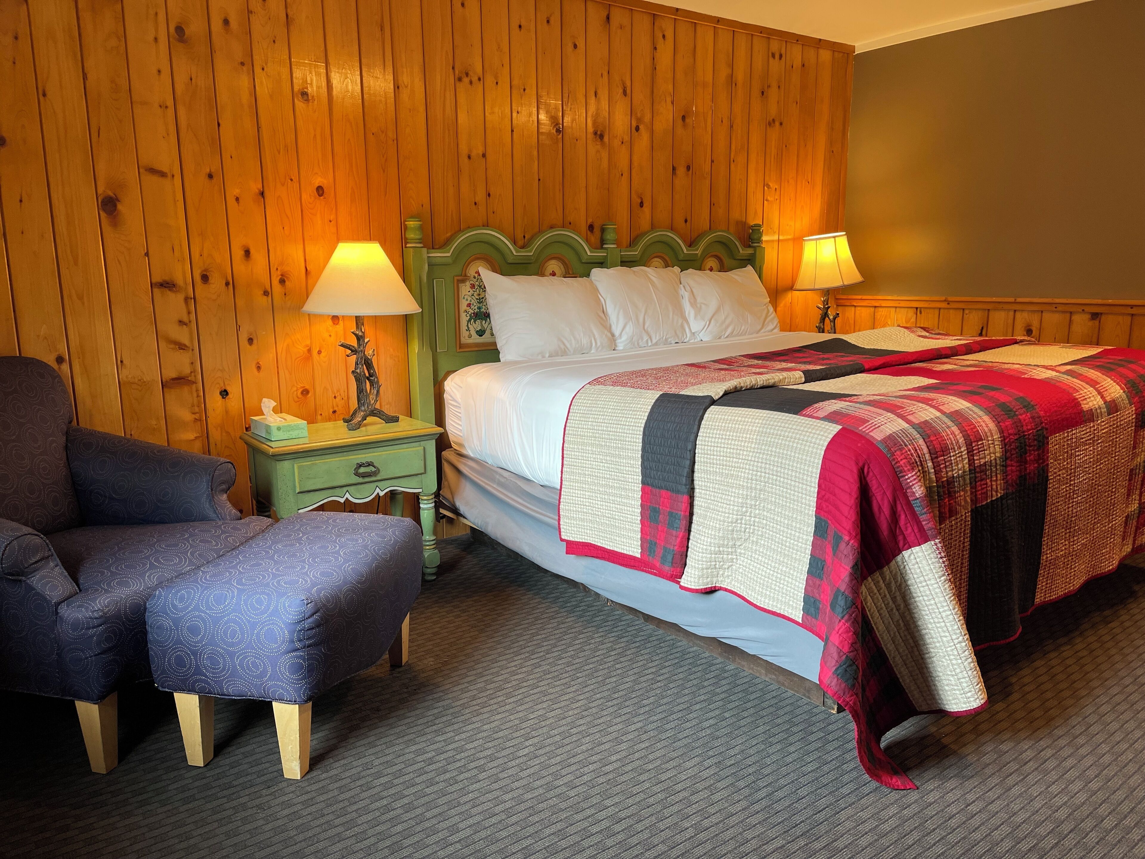 Una habitación con pared de paneles de madera, una cama con cabecera verde, dos mesitas de noche con lámparas, una silla y un reposapiés estampado.
