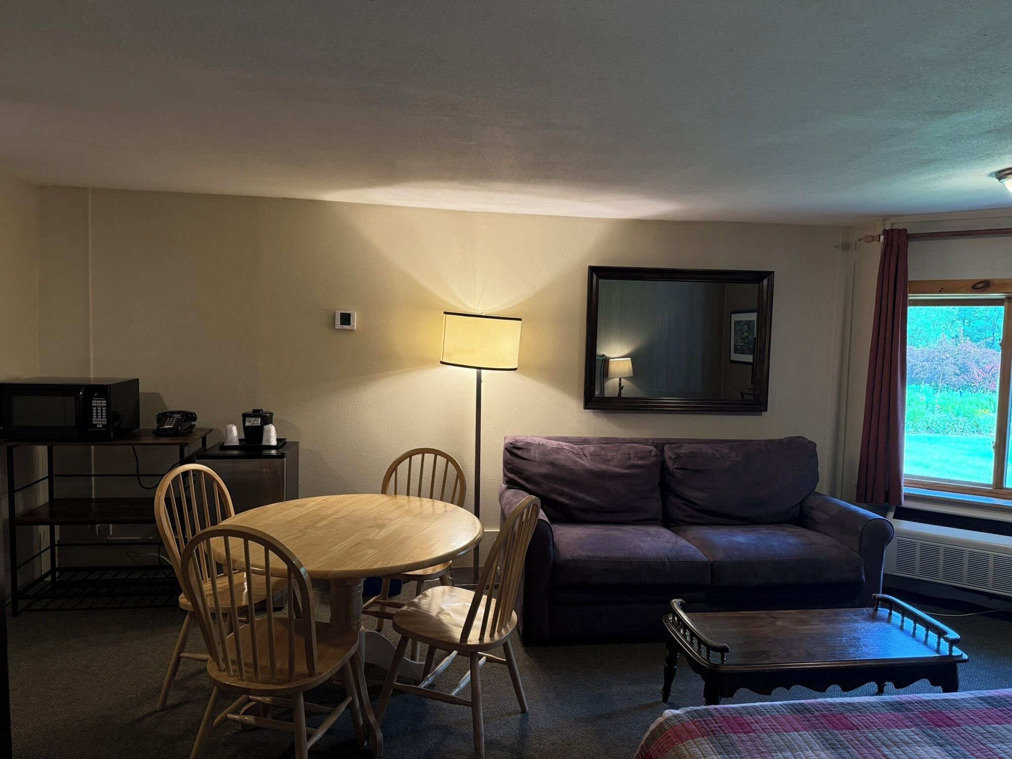 A room with a round wooden table, four wooden chairs, a dark-colored sofa, a small wooden coffee table, a lamp, a television, and a window with curtains.