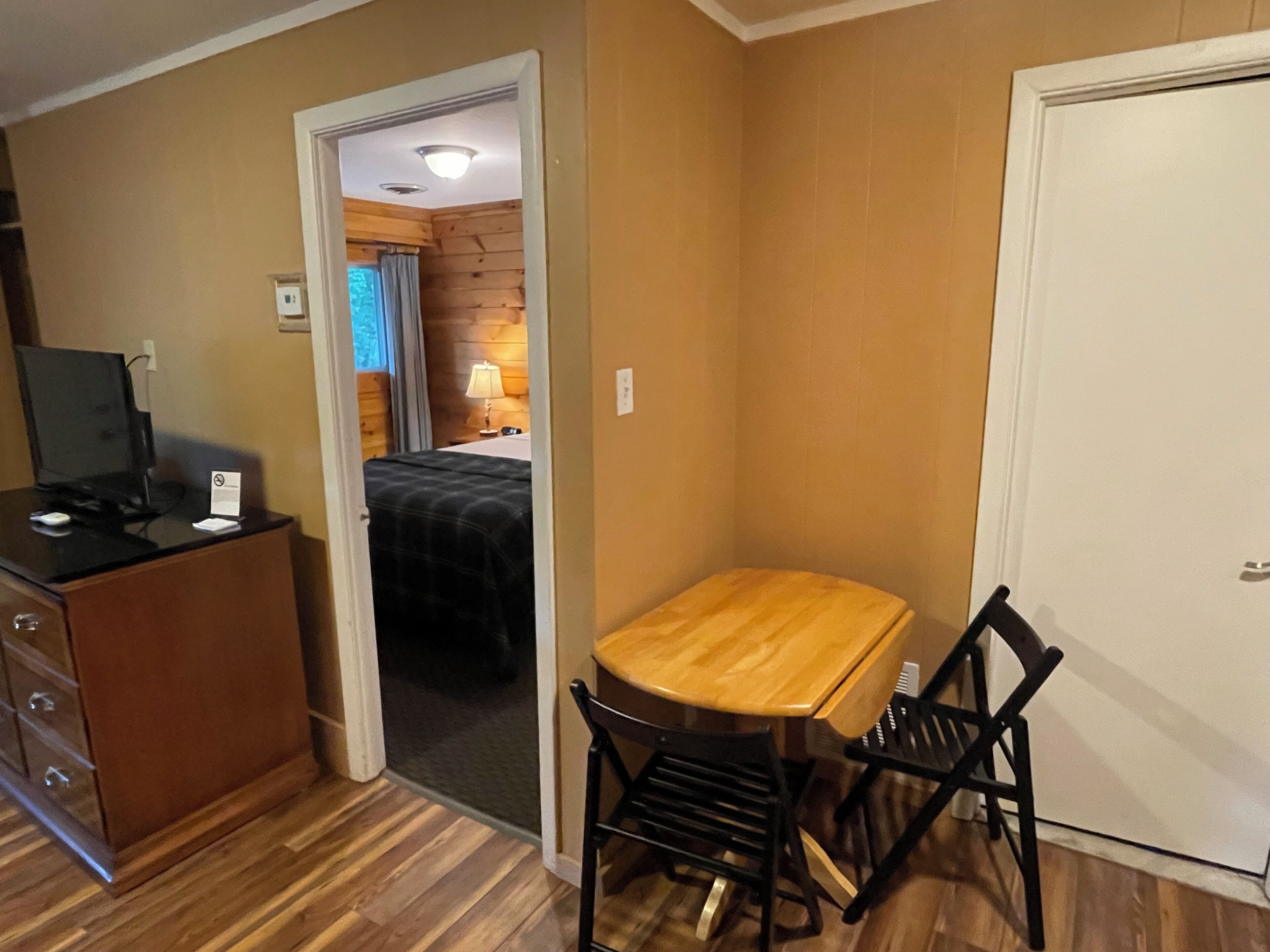 A compact room with a wooden desk, two chairs, a small cabinet, and a bed in the adjacent room.