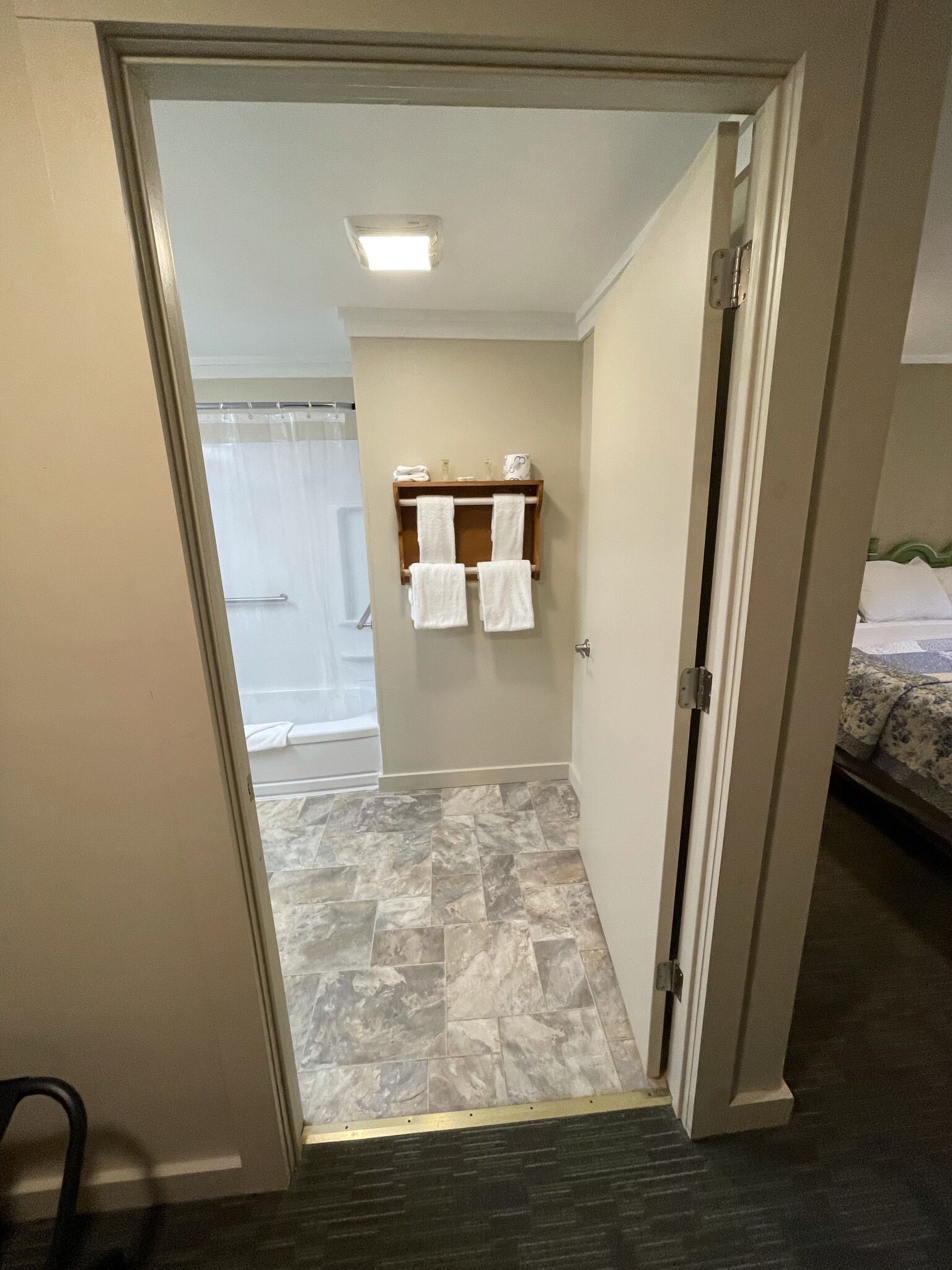 A hotel bathroom with a shower, a towel rack with towels, and a bed with a floral bedspread.