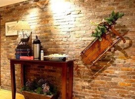 Pared de ladrillo rústico con un perchero de madera que sostiene una planta y un adorno. Frente a la pared, una mesa con libros, una lámpara y un termo.