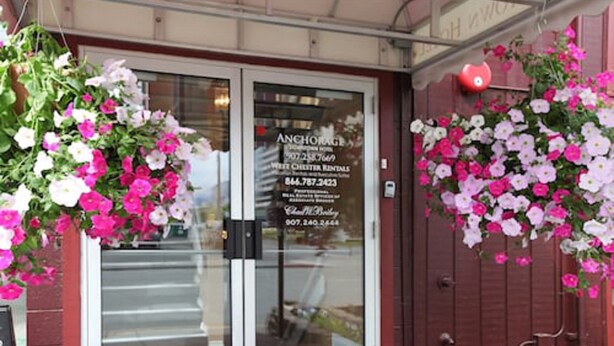 A hotel entrance with a glass door, a signboard with contact information, and hanging flower arrangements. - Anchorage Downtown Hotel (Anchorage)
