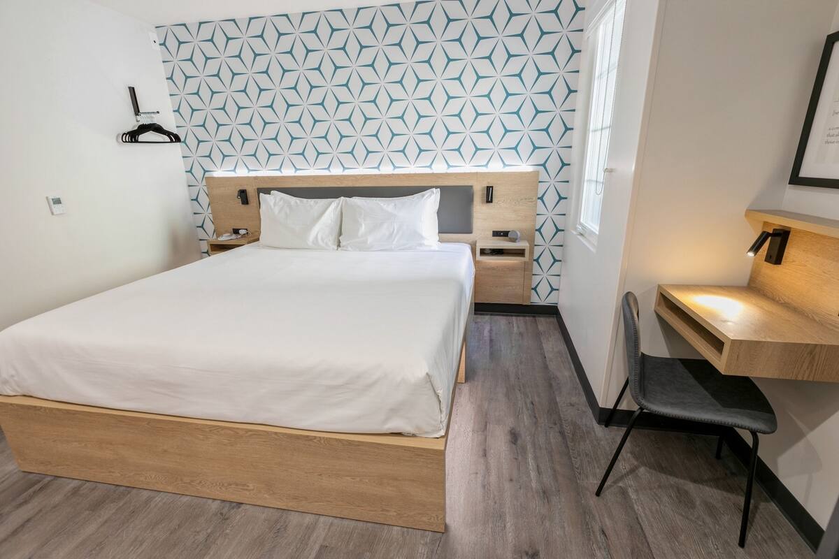 A modern hotel room with a wooden bed, a desk with a chair, and a geometric patterned wall.