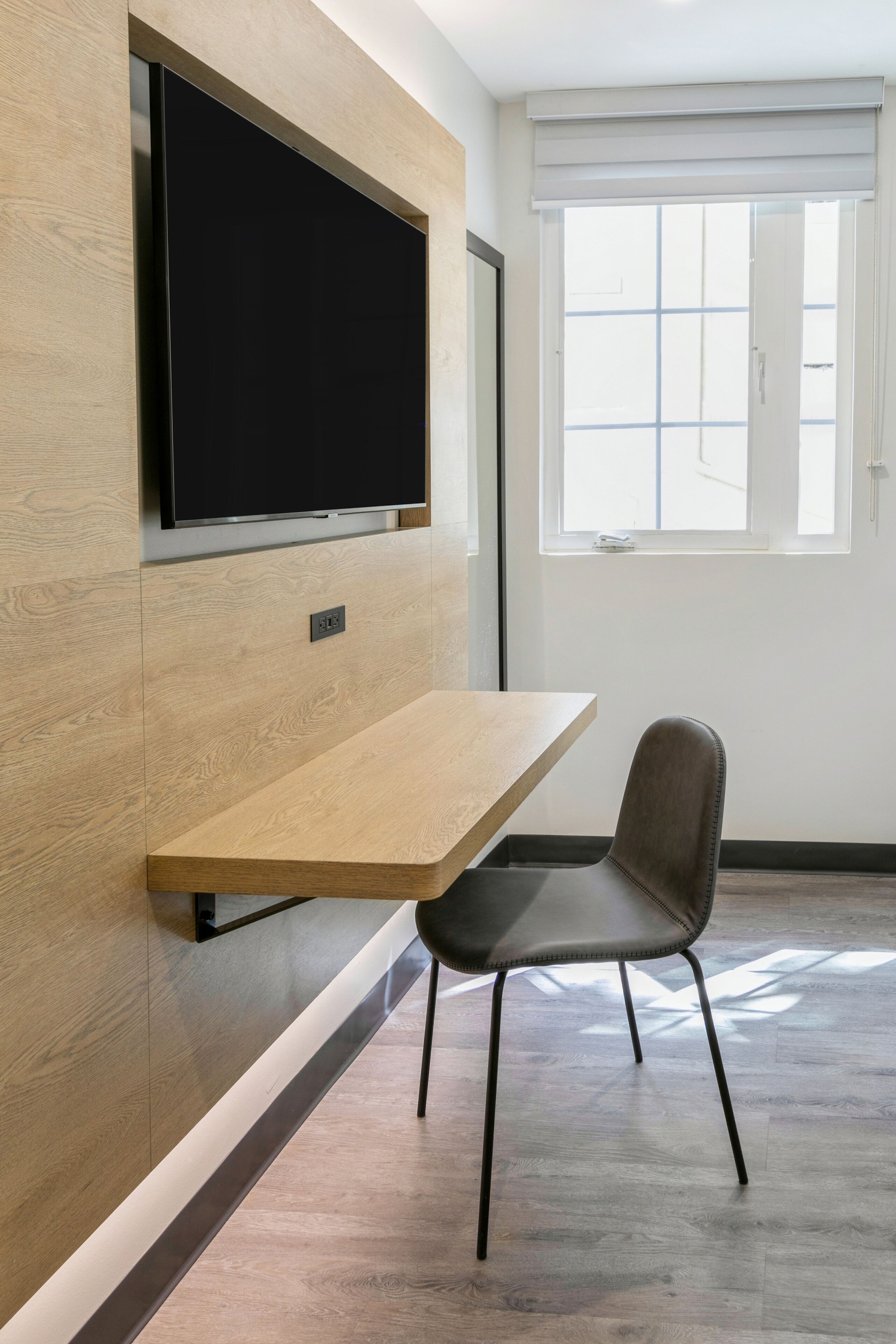 A modern room with a built-in TV, a wooden desk, a chair, and a window with blinds.