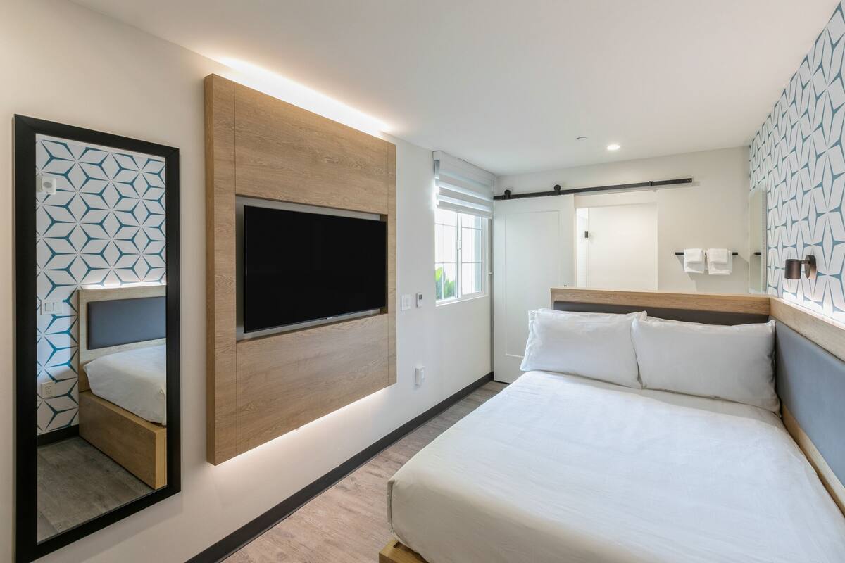A modern hotel room with a large bed, a mounted TV, and a mirror reflecting the room.